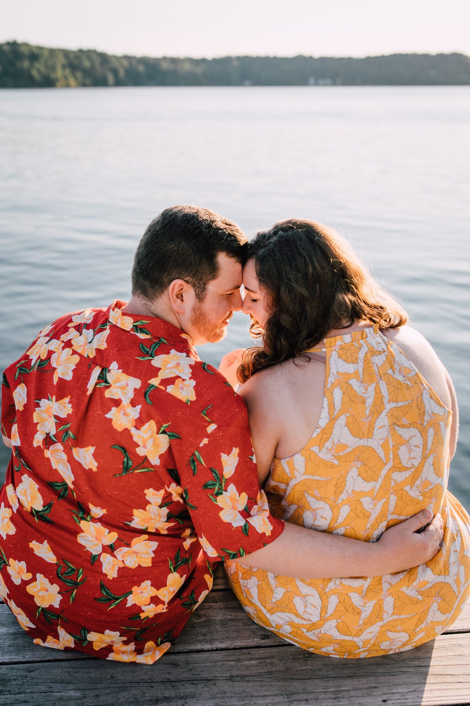  Engaged couple lean their faces in close to one another while sitting on a dock in front of Lake Ontario for their sunset engagement photos 