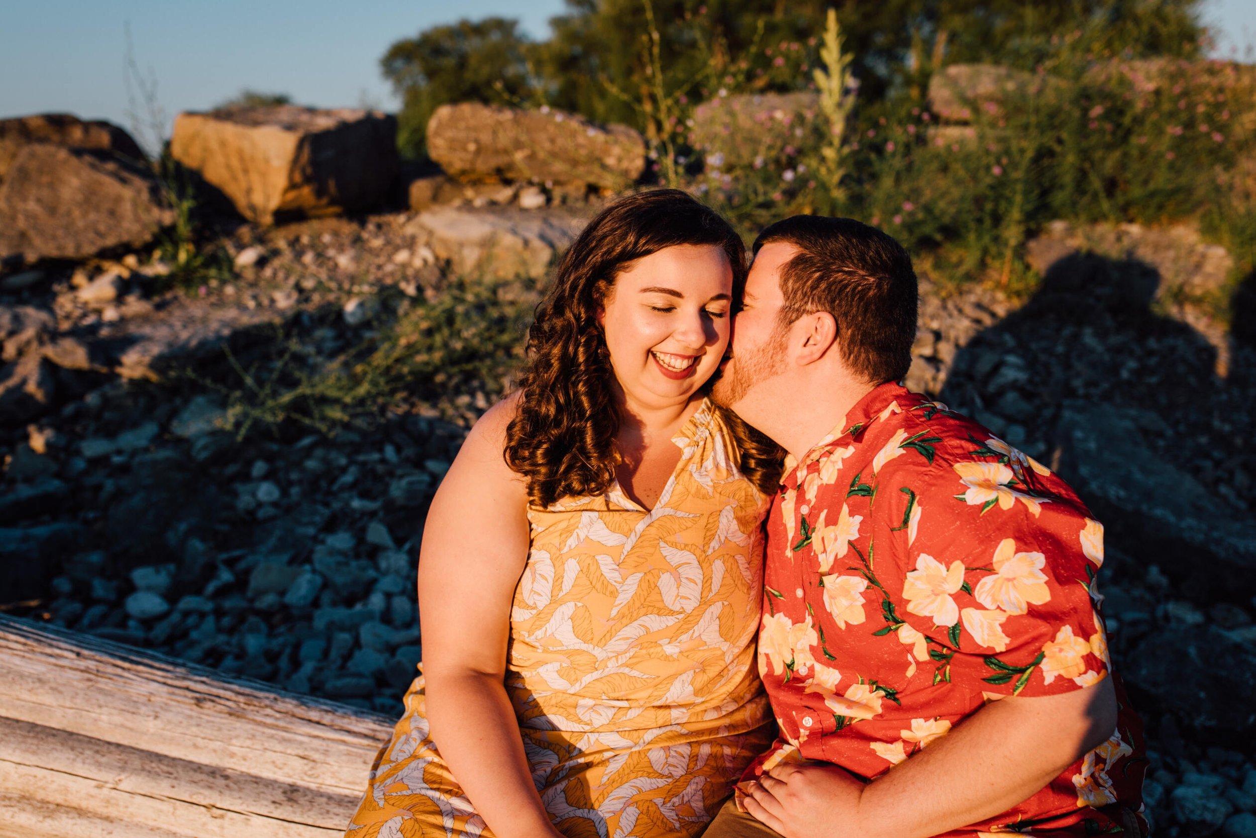  Brandon kisses his fiancé’s cheek as they sit on a log on a rocky beach for sunset engagement photos 