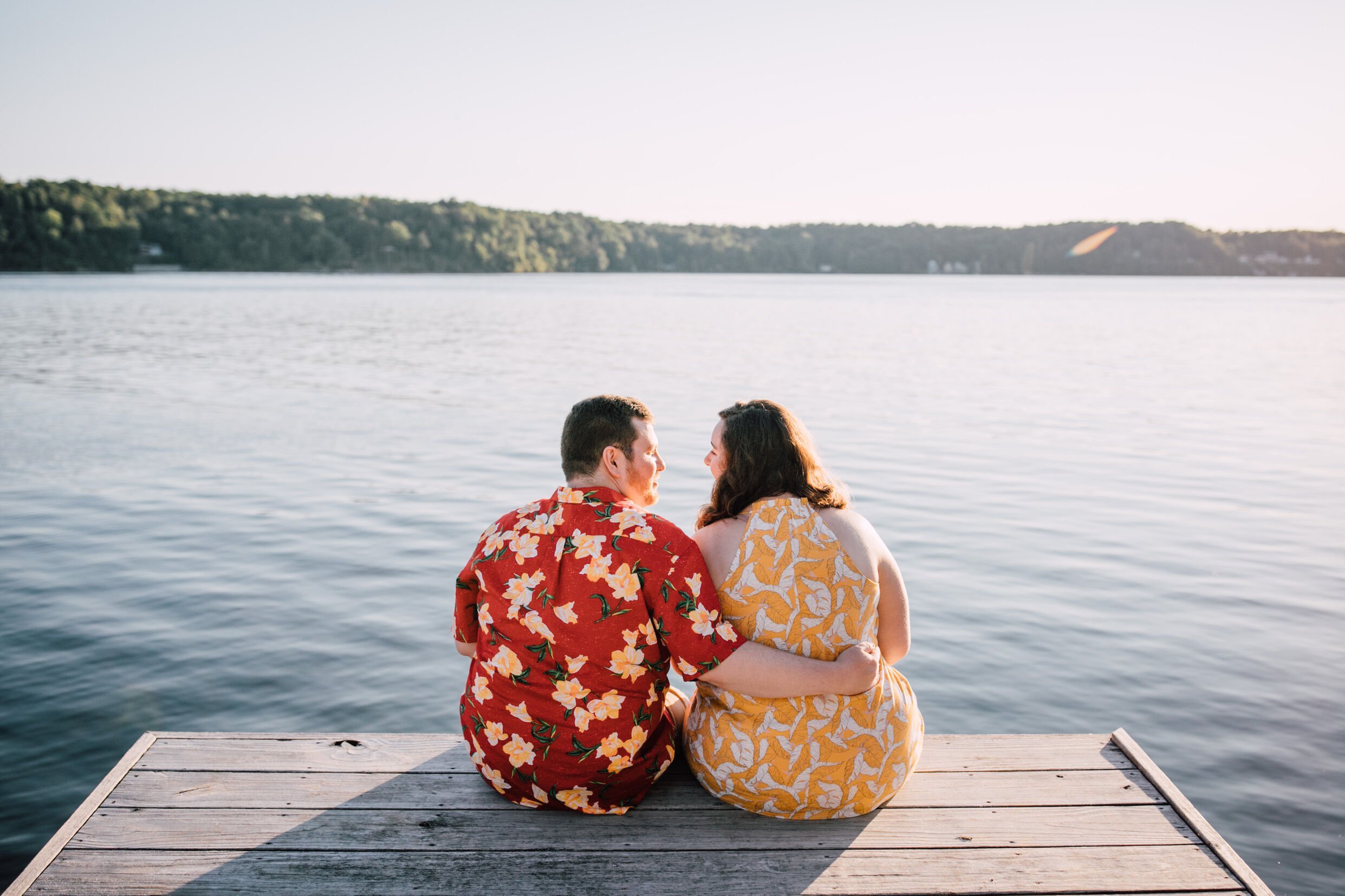  An engaged couple sit together at the end of a wooden dock on the coast of Lake Ontario and look at each other for sunset engagement photos with the water in front of them 