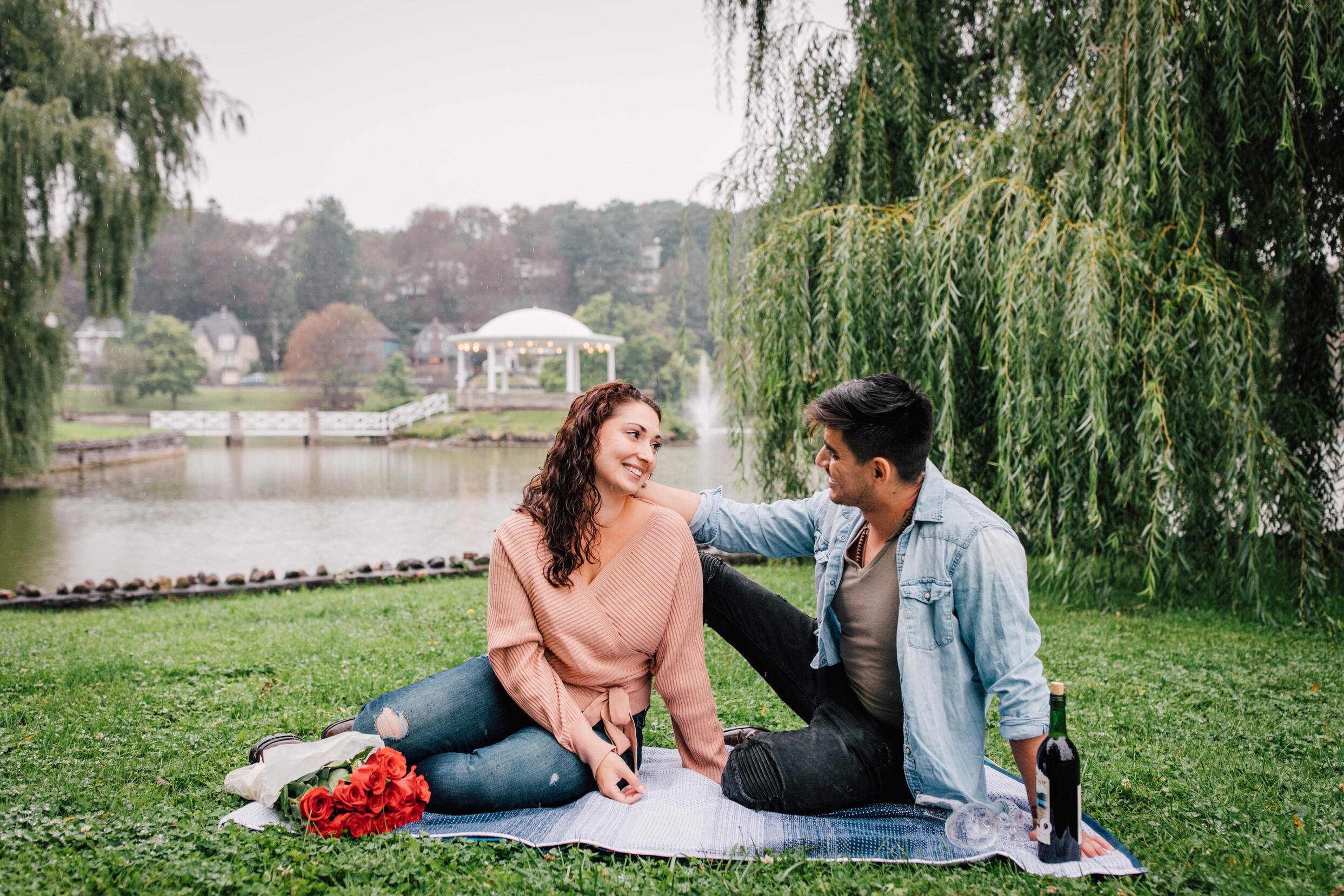  jessica and pablo smile at each other as they sit on a picnic blanket in a grassy area surrounded by the willow trees in upper onondaga park in front of a lake 