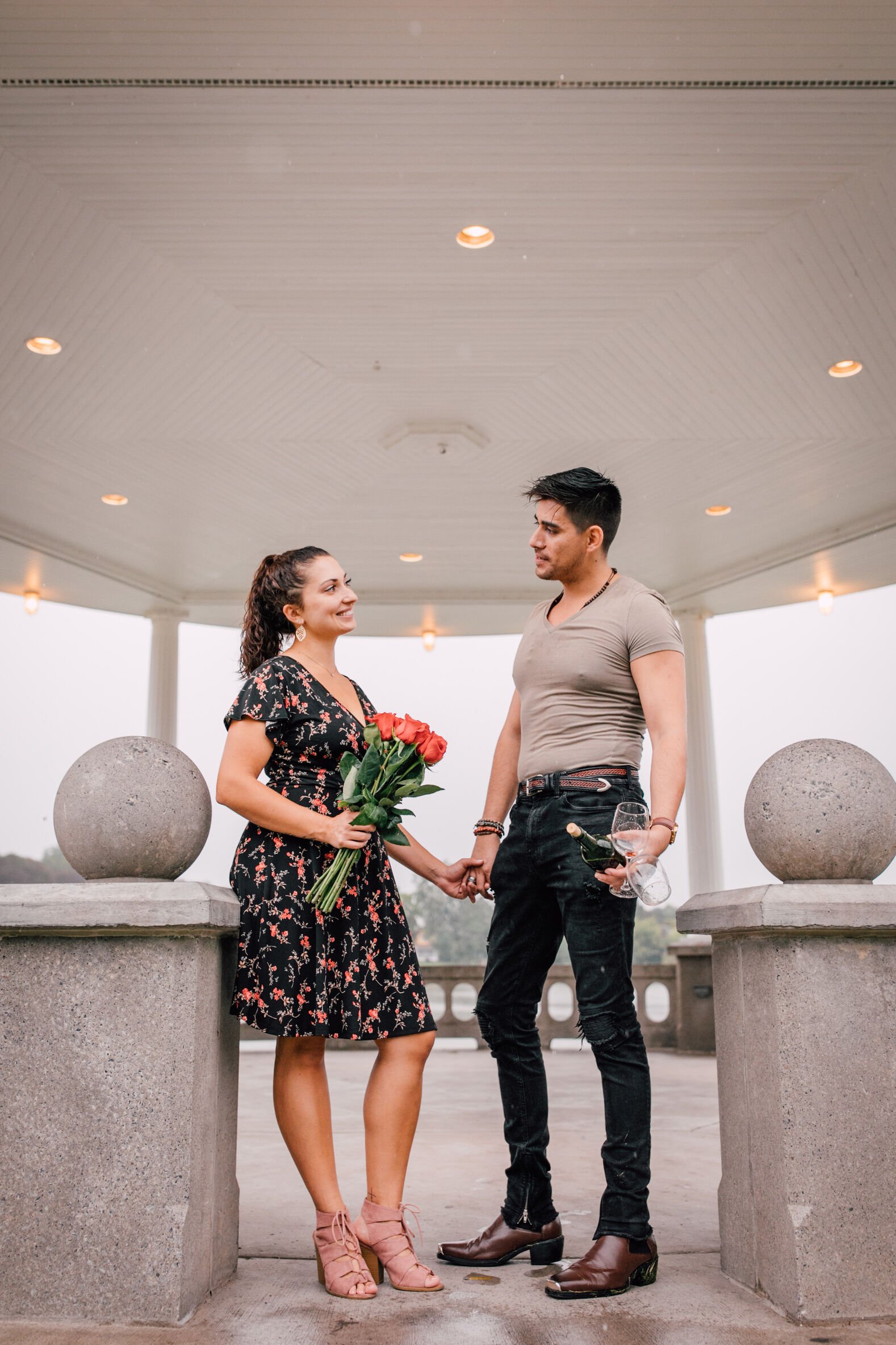  pablo and jessica hold hands while he holds a wine bottle and two glasses and she holds a bouquet of roses during their engagement photos in the rain 