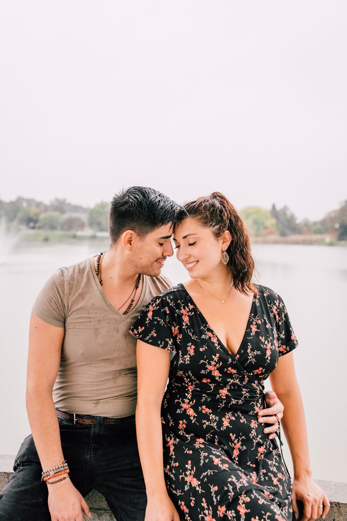  pablo and jessica lean into each other as they smile while sitting on the ledge of a gazebo in front of a lake during their engagement photos in the rain 