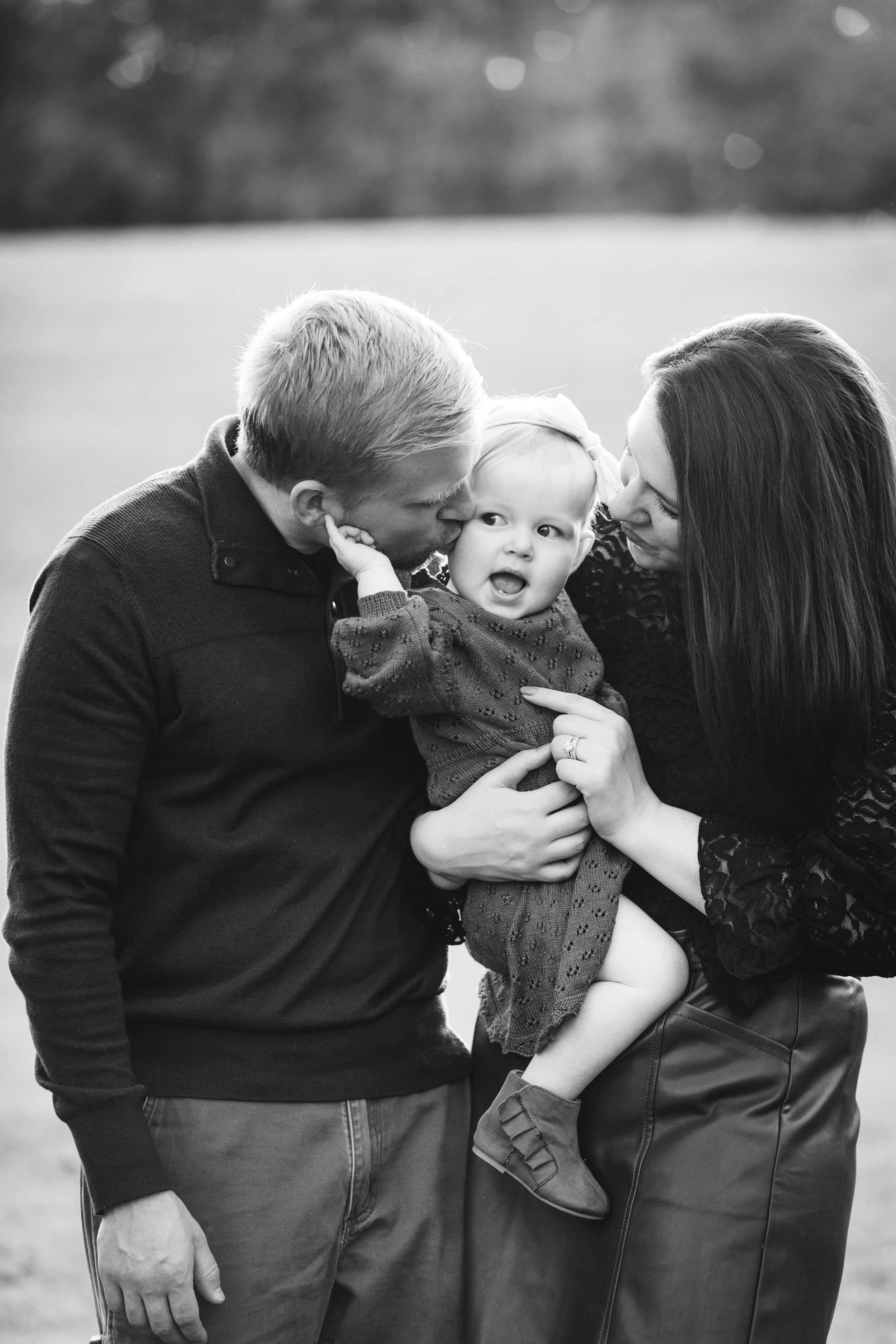  dad kisses baby girl’s cheek as mom holds and smiles at her during extended family portraits 