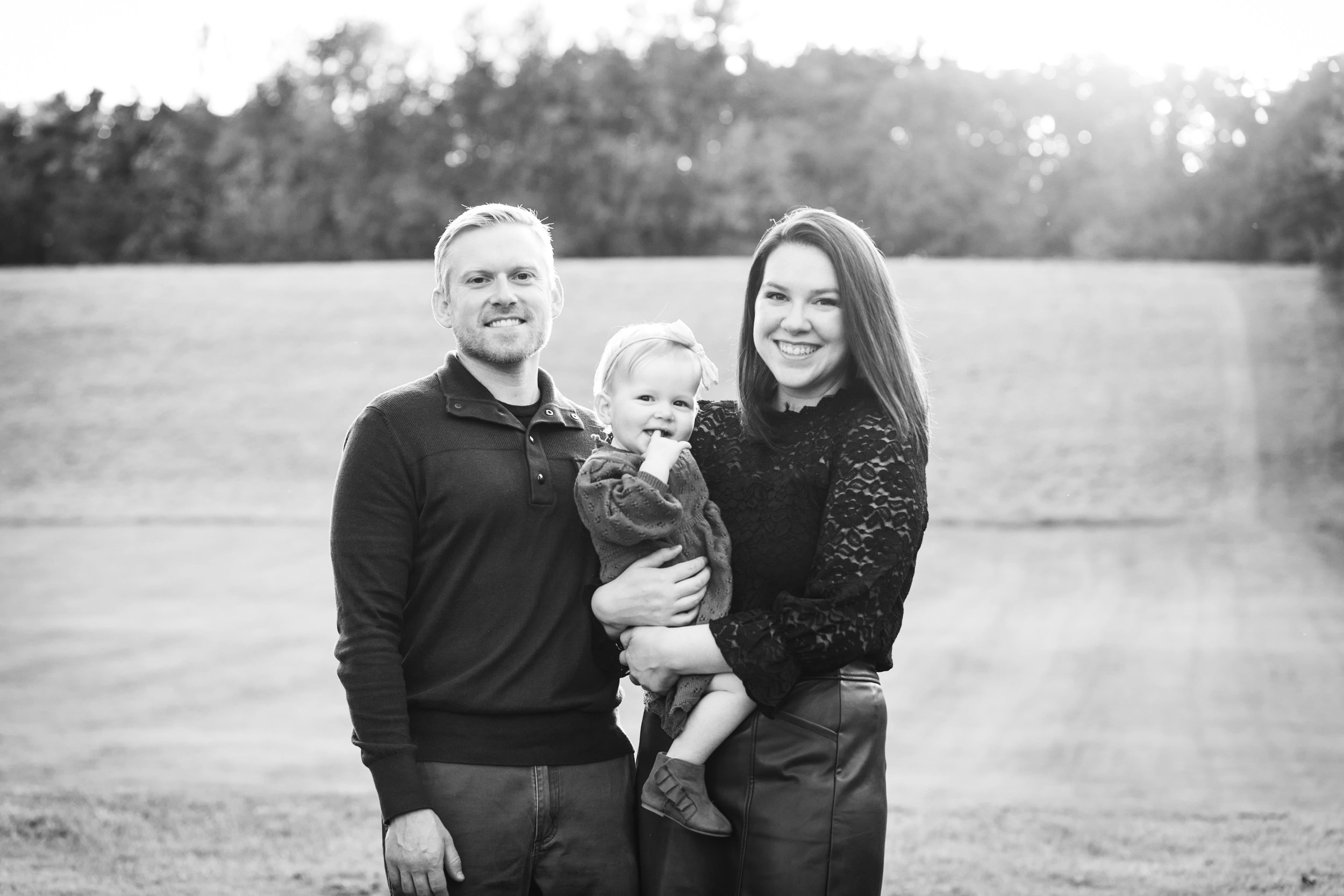 mom dad and baby girl smile as they stand in front of an open field as the sun sets during fall family photos 