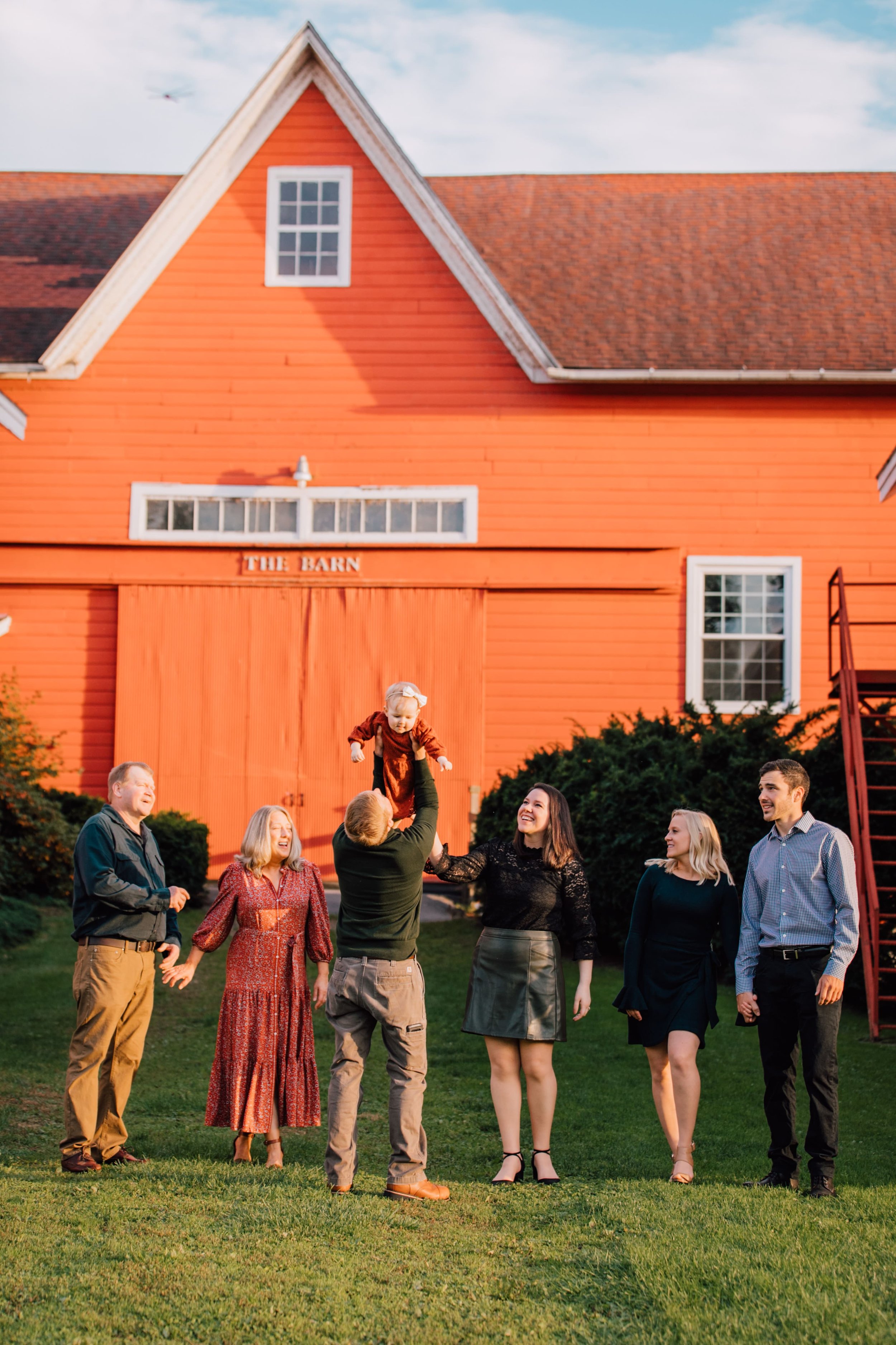  family of 7 look at baby girl as dad lifts her up they stand in front of a red barn during extended family photos 