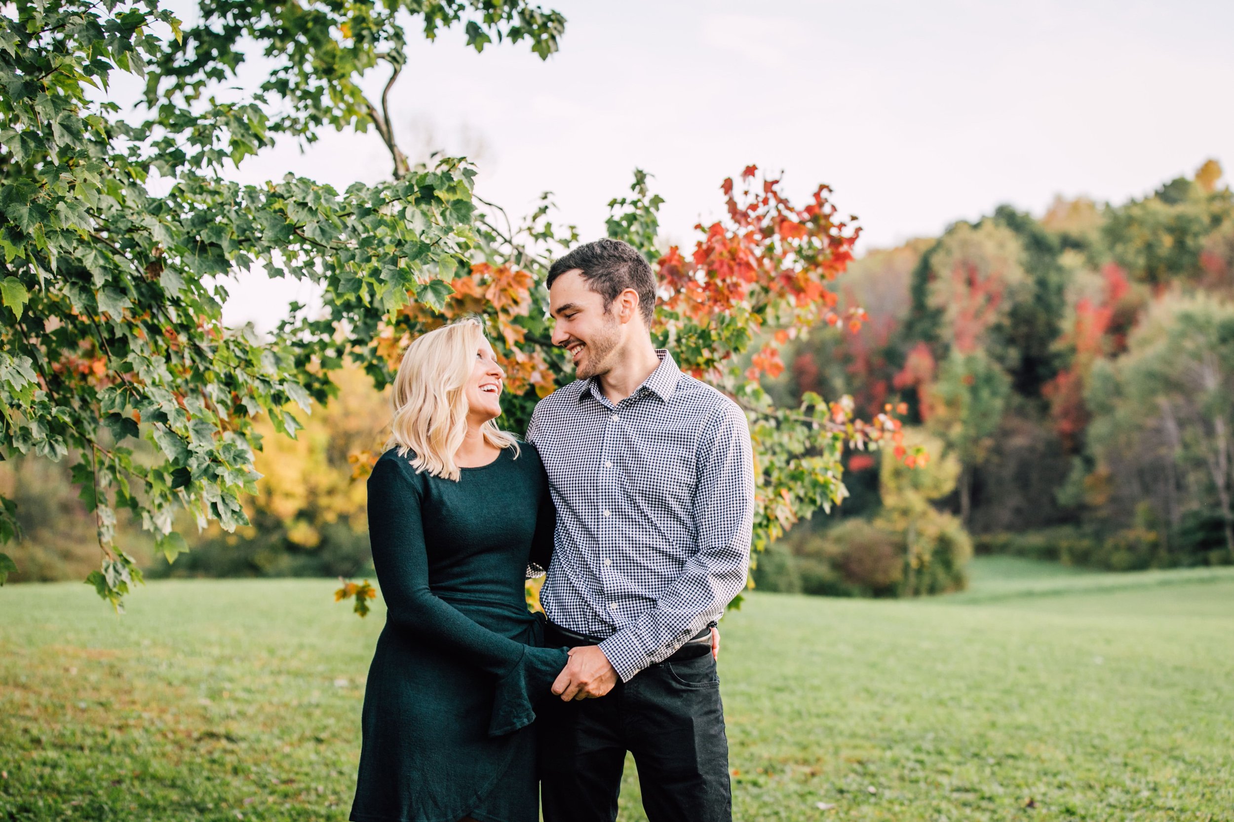  engaged couple smile at each other while holding hands as they stand in front of a tree that is changing colors for fall extended family photos 