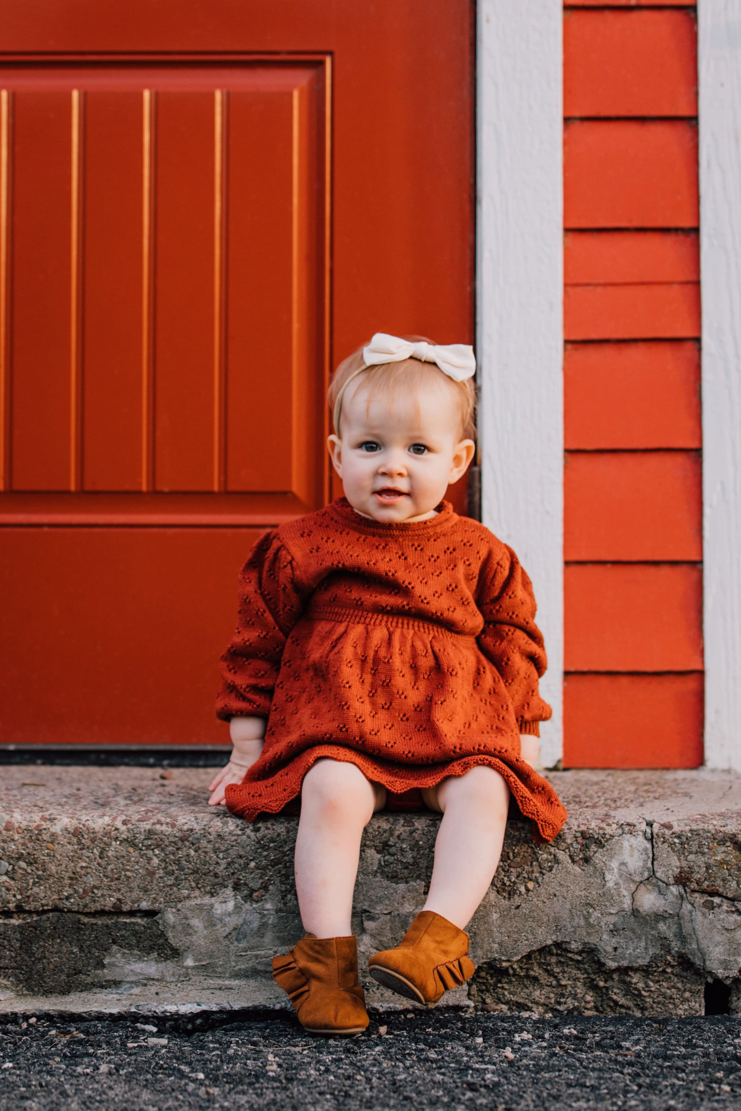  a baby girl sits on a step in front of a red barn and smiles as she wears an orange dress and a white bow in her hair during family photos in Oswego, NY 