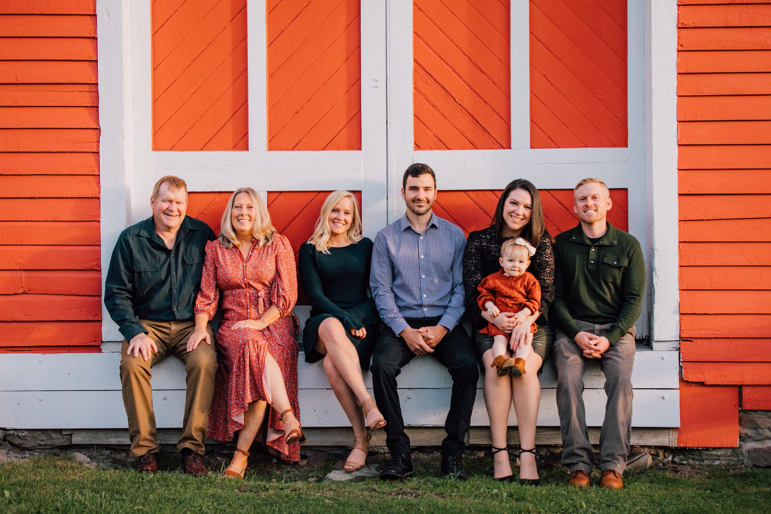  a family of 7 sit in front of a red barn wearing fall attire during extended family photos 