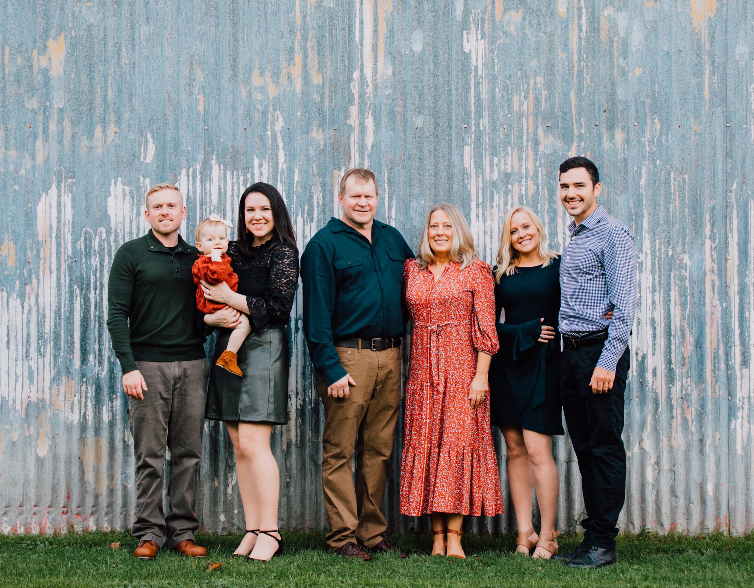  a family of 7 stand in fall attire in front of a blue rustic looking wall during adult family photos 