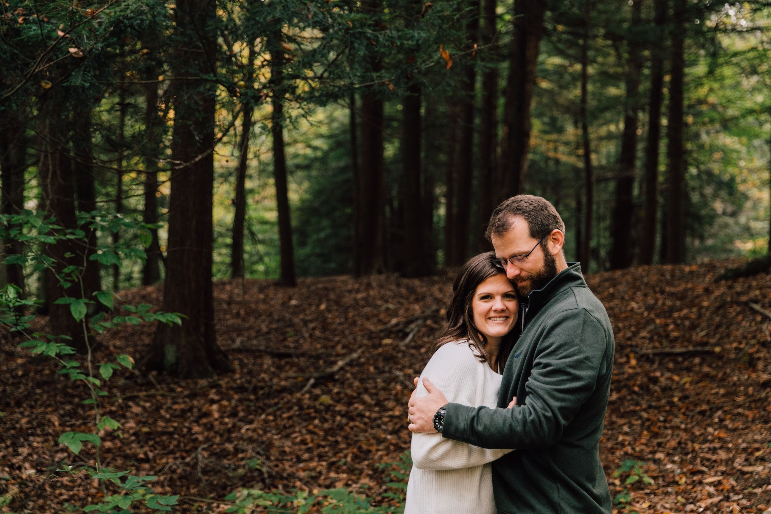  megan and barrett stand together on their wooded property in an embrace megan looks at the camera and smiles while barrett presses his face to her forehead with closed eyes during their couples photoshoot 
