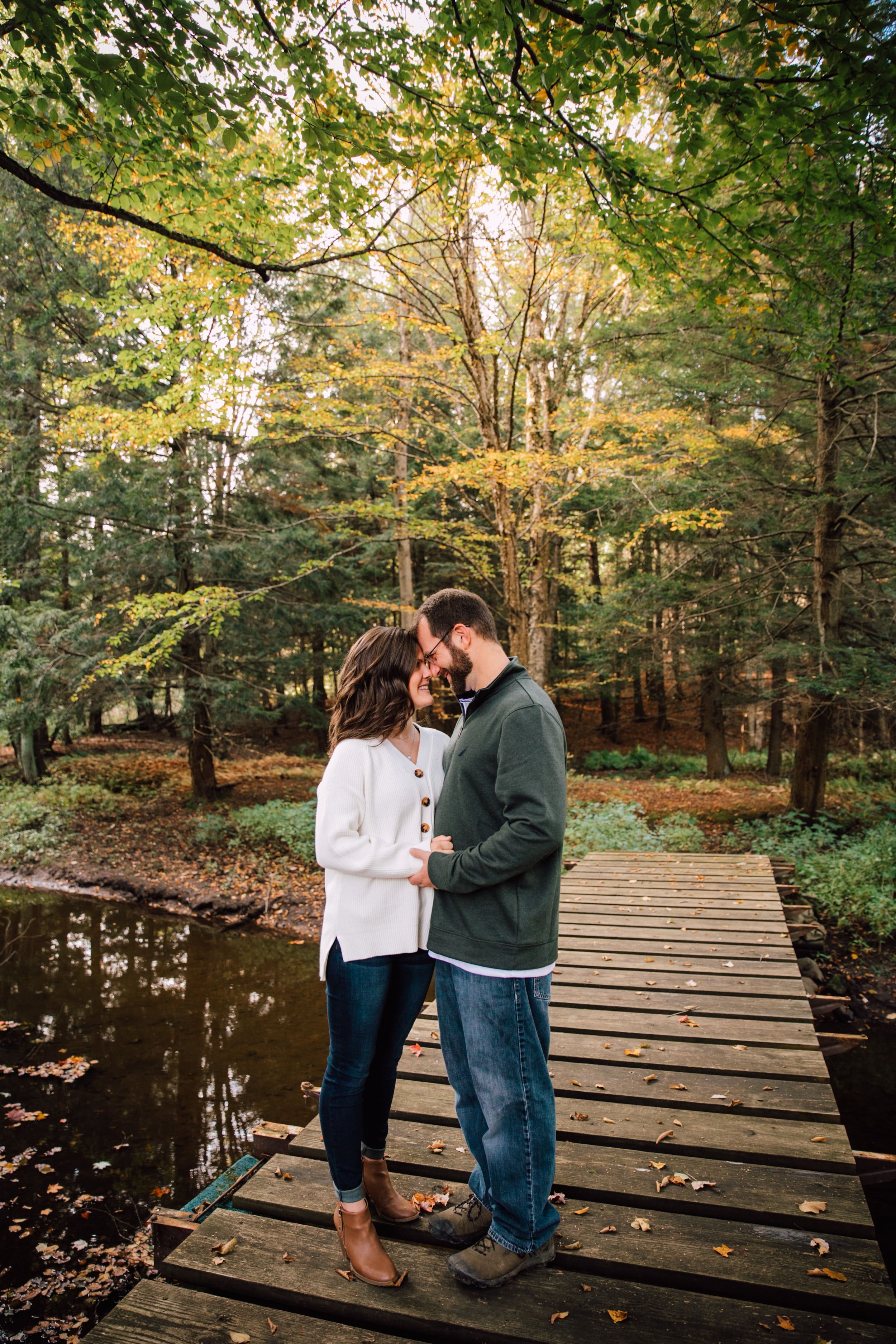  megan and barrett stand on a small bridge over a creek during their forest engagement photos and press their foreheads together while smiling at each other 