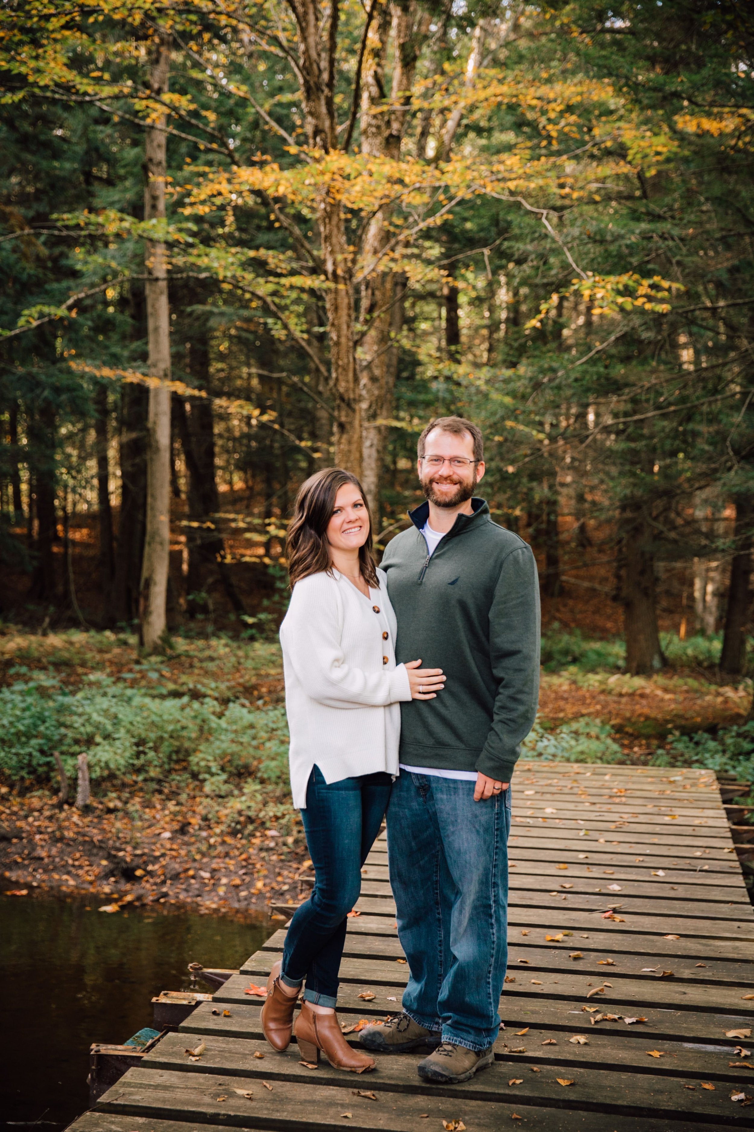  megan and barrett smile as they stand on a wooded bridge in the wooded area on their property during a backyard photoshoot to celebrate their wedding 