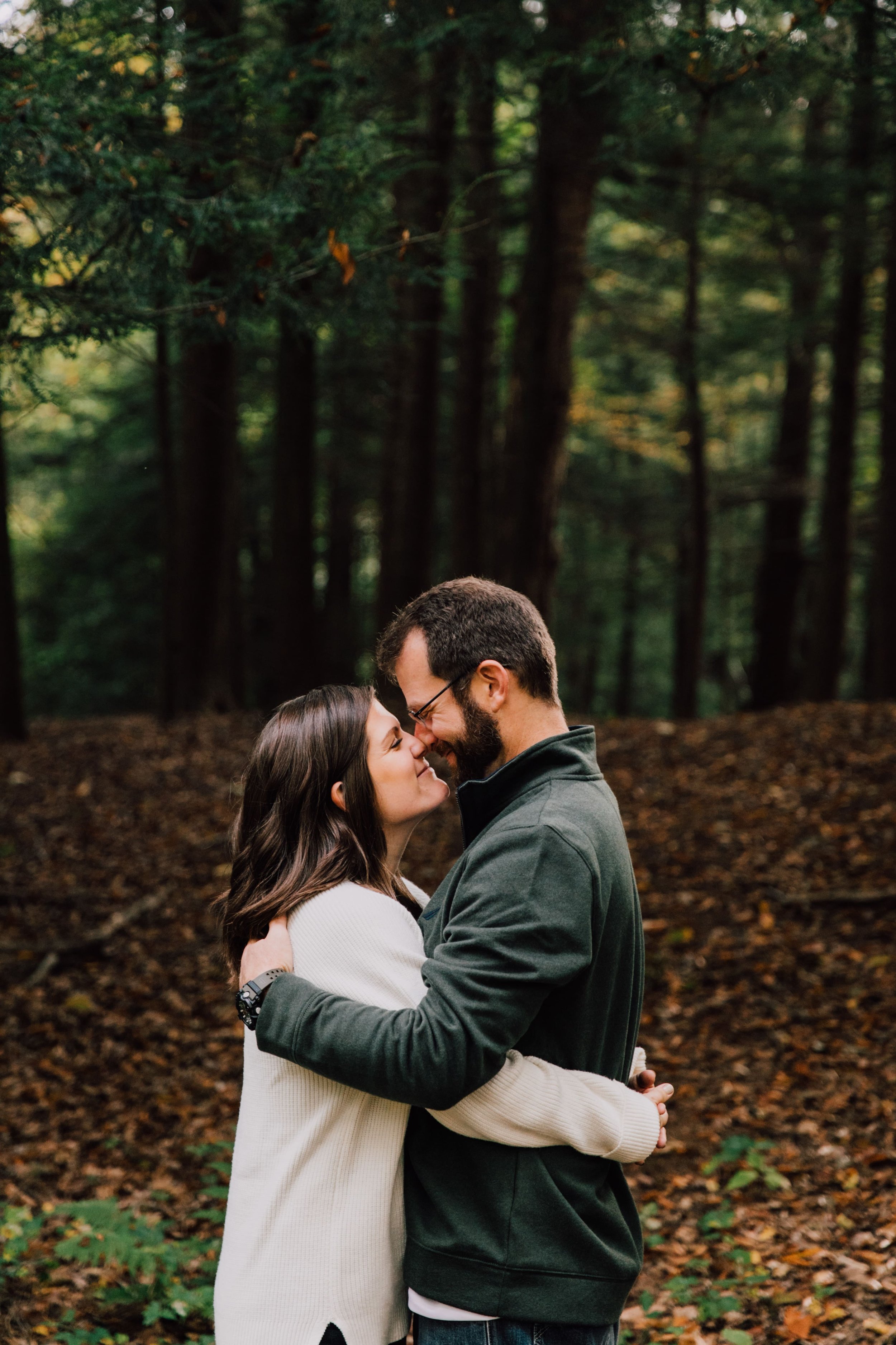  megan and barrett stand for forest engagement photos with their noses pressed together as they smile in an embrace in the wooded area in their backyard 