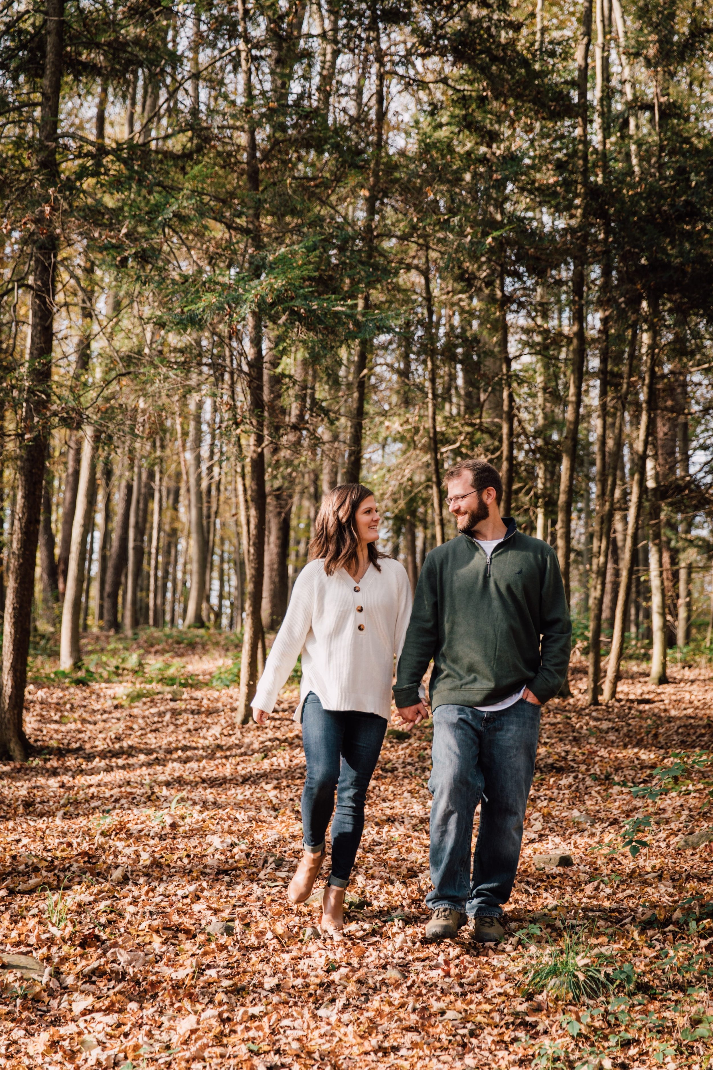  megan and barrett walk hand in hand around their forest property while wearing fall sweaters and jeans during their backyard photoshoot 