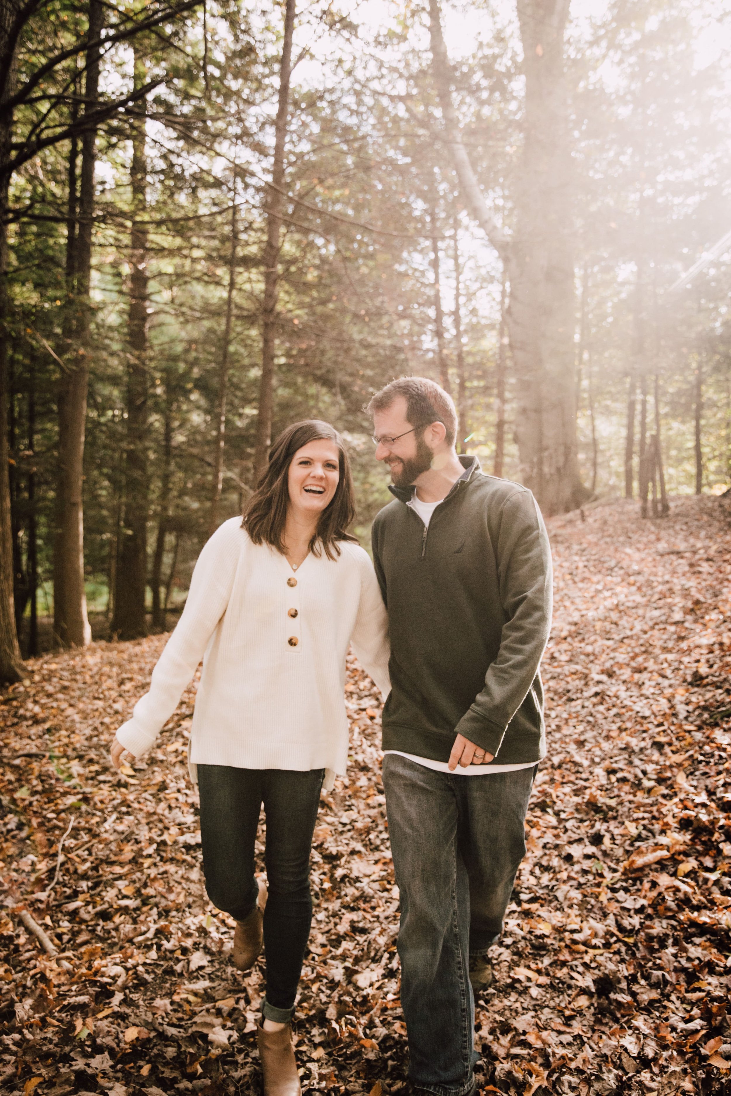  megan and barrett’s fall engagement photos to celebrate their wedding they walk together through a wooded area while smiling megan wears a white waffle knit sweater with jean and brown boots and barrett wears an olive green quarter zip with jeans an