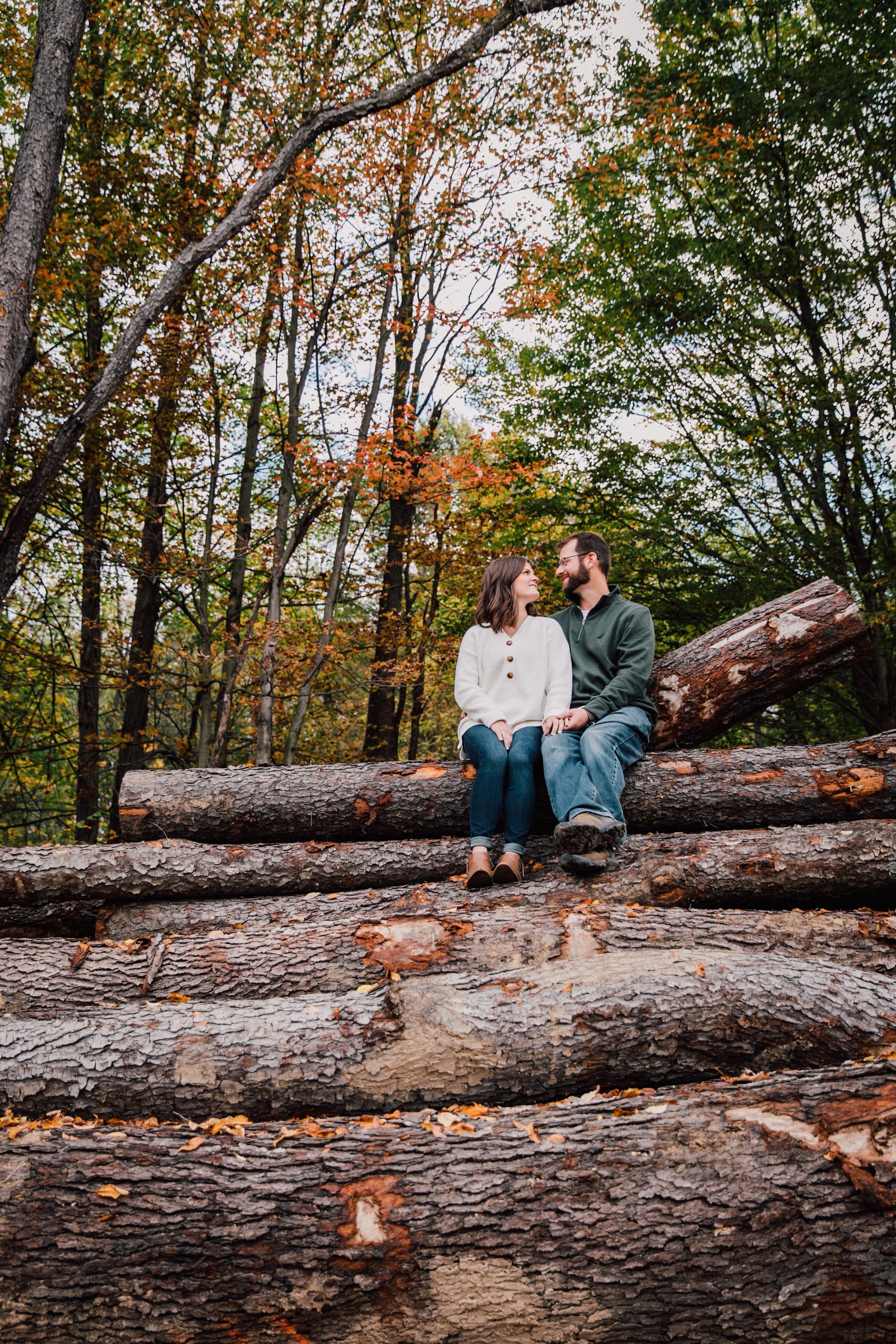  megan and barrett sit atop a pile of logs during their forest engagement photos session to celebrate their marriage the sit looking at each other while smiling and wearing fall outfits 