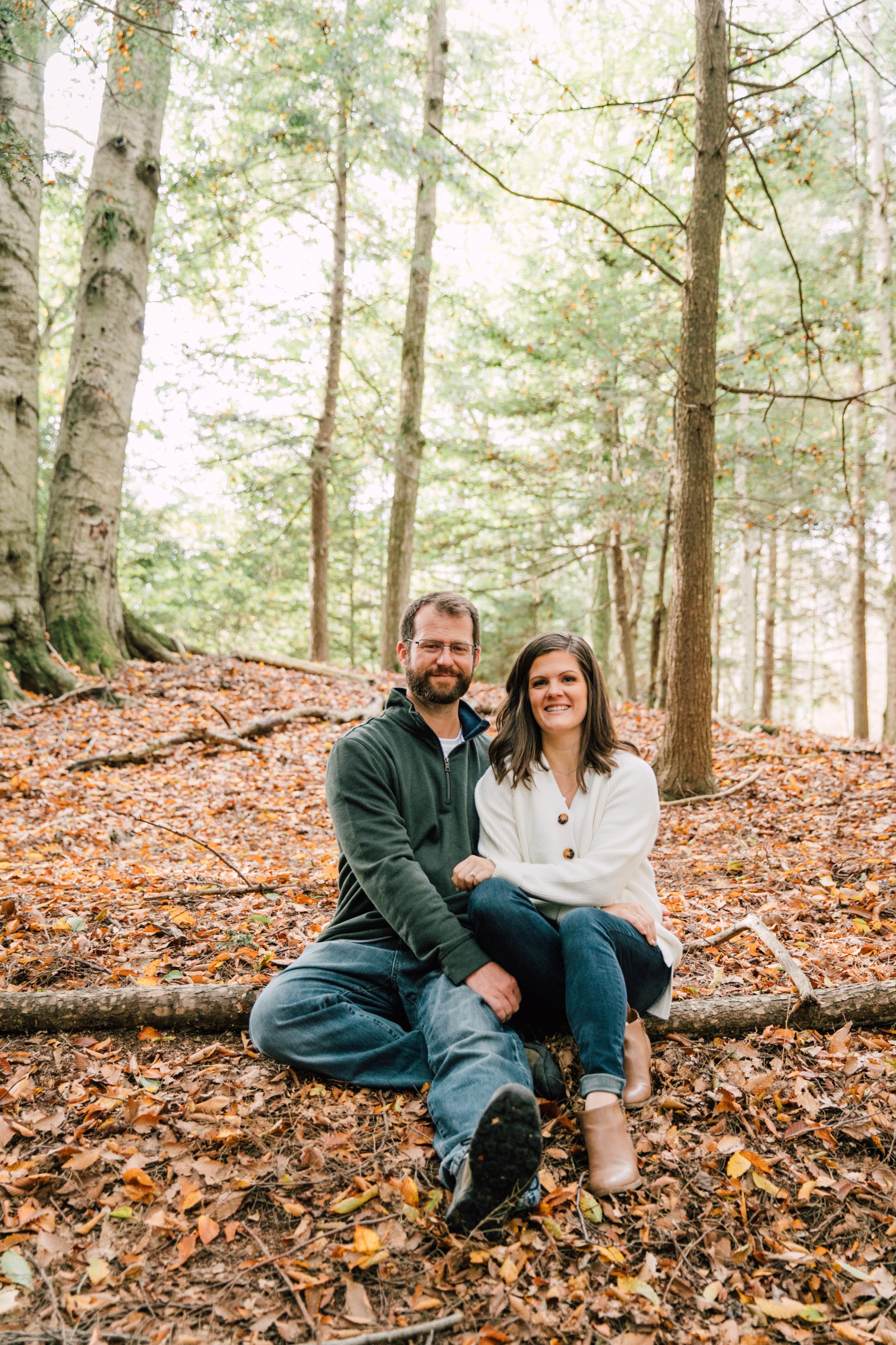  the couple sit next to each other on the fall leaf lined ground in front of a log and smile as they cuddle up during their fall engagement photos 
