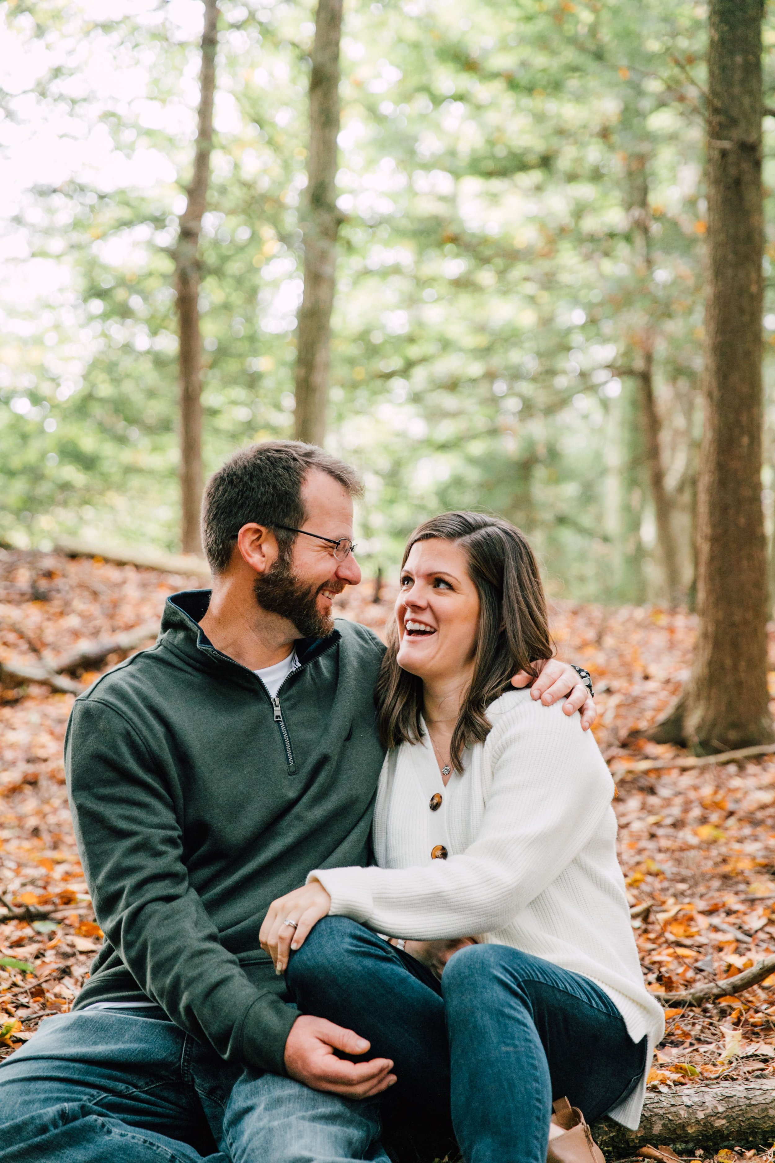  the couple laughs together as they cuddle up on the fall leaf lined ground during their forest engagement photos 