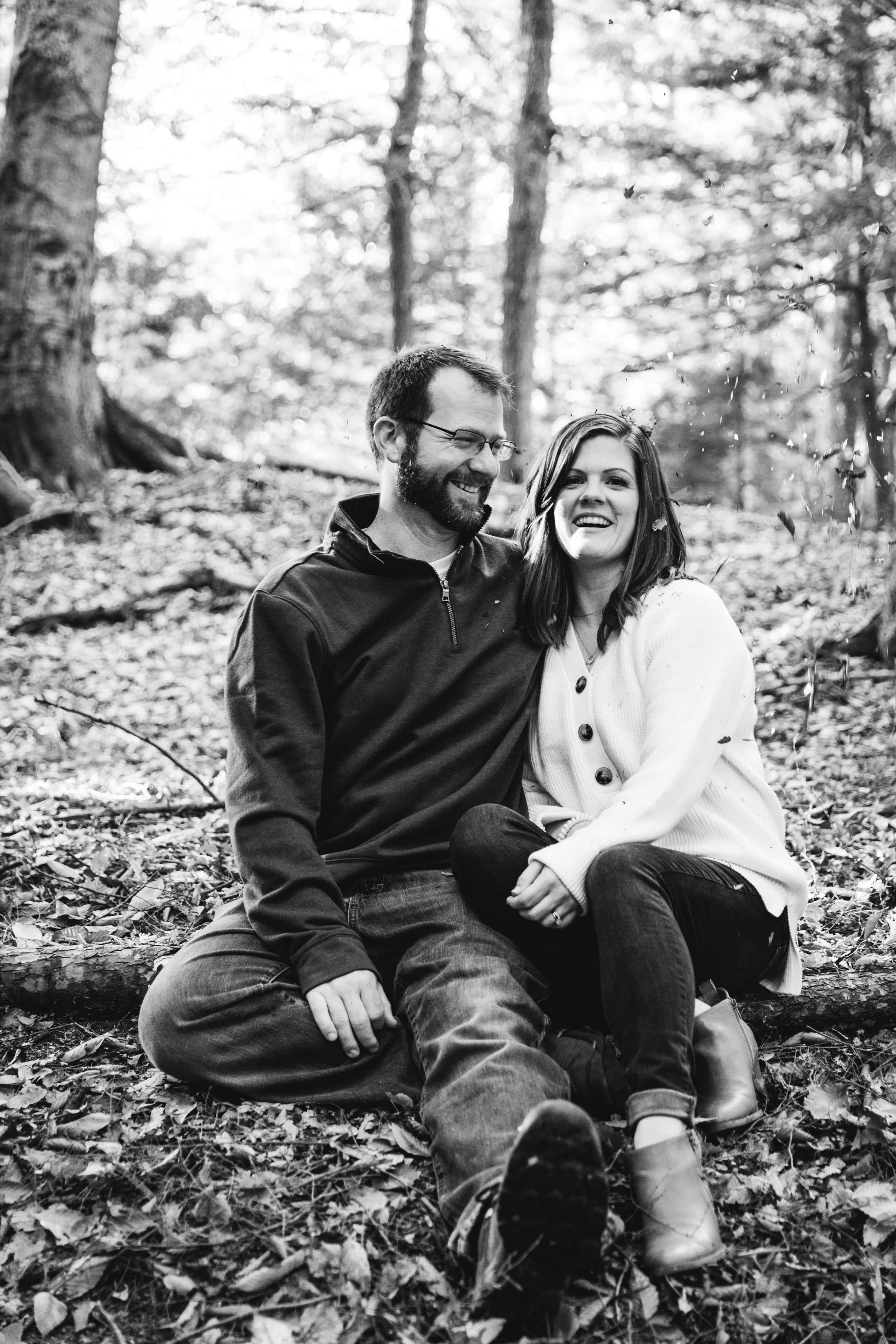  the couple laugh as fall leaves fall around them while they sit on the ground during their forest engagement session 