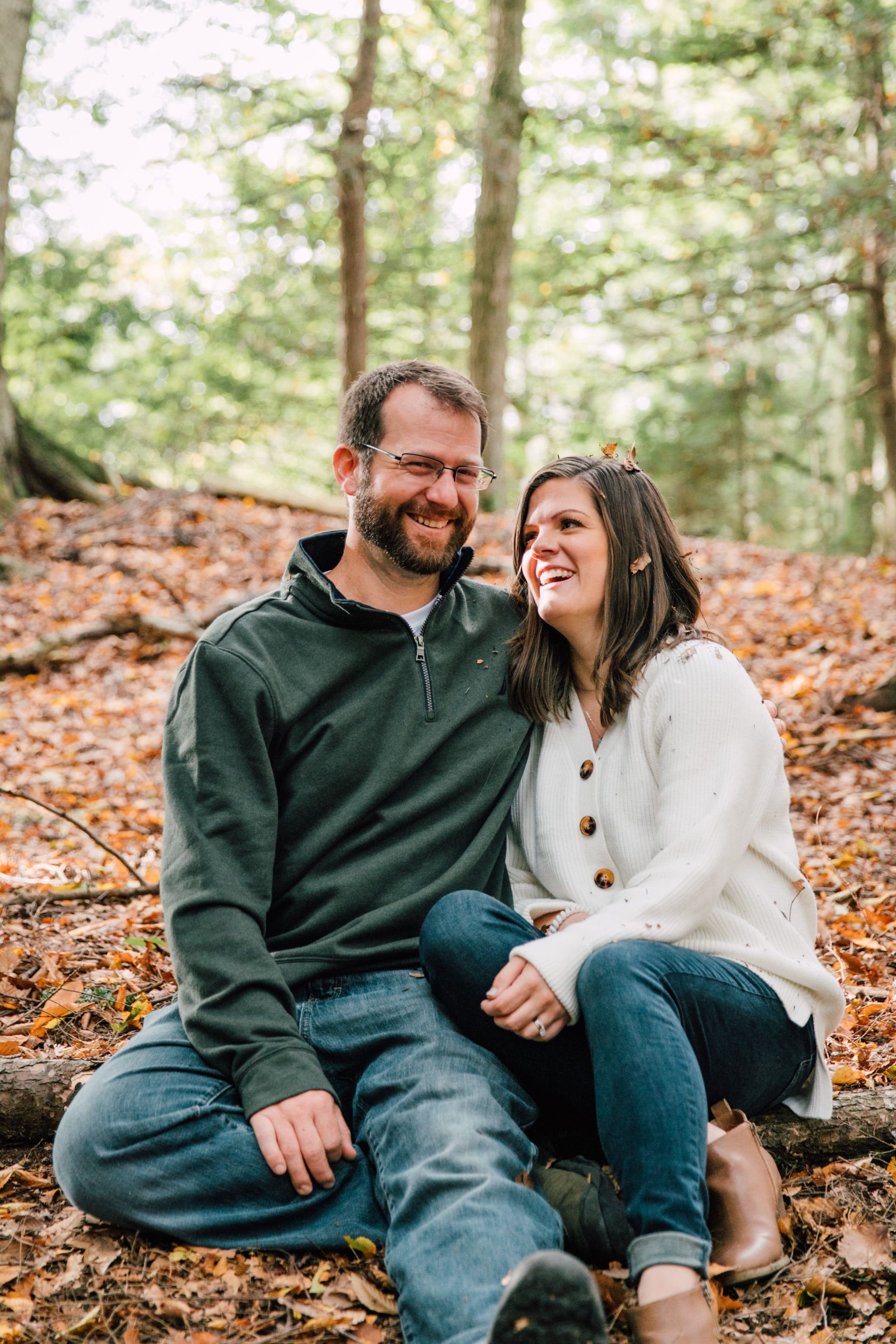  megan and barret’s fall engagement photos are full of laughs and leaves pictured here, they laugh together as they are cuddled up on the fall leaf lined ground with falling leaves around them 