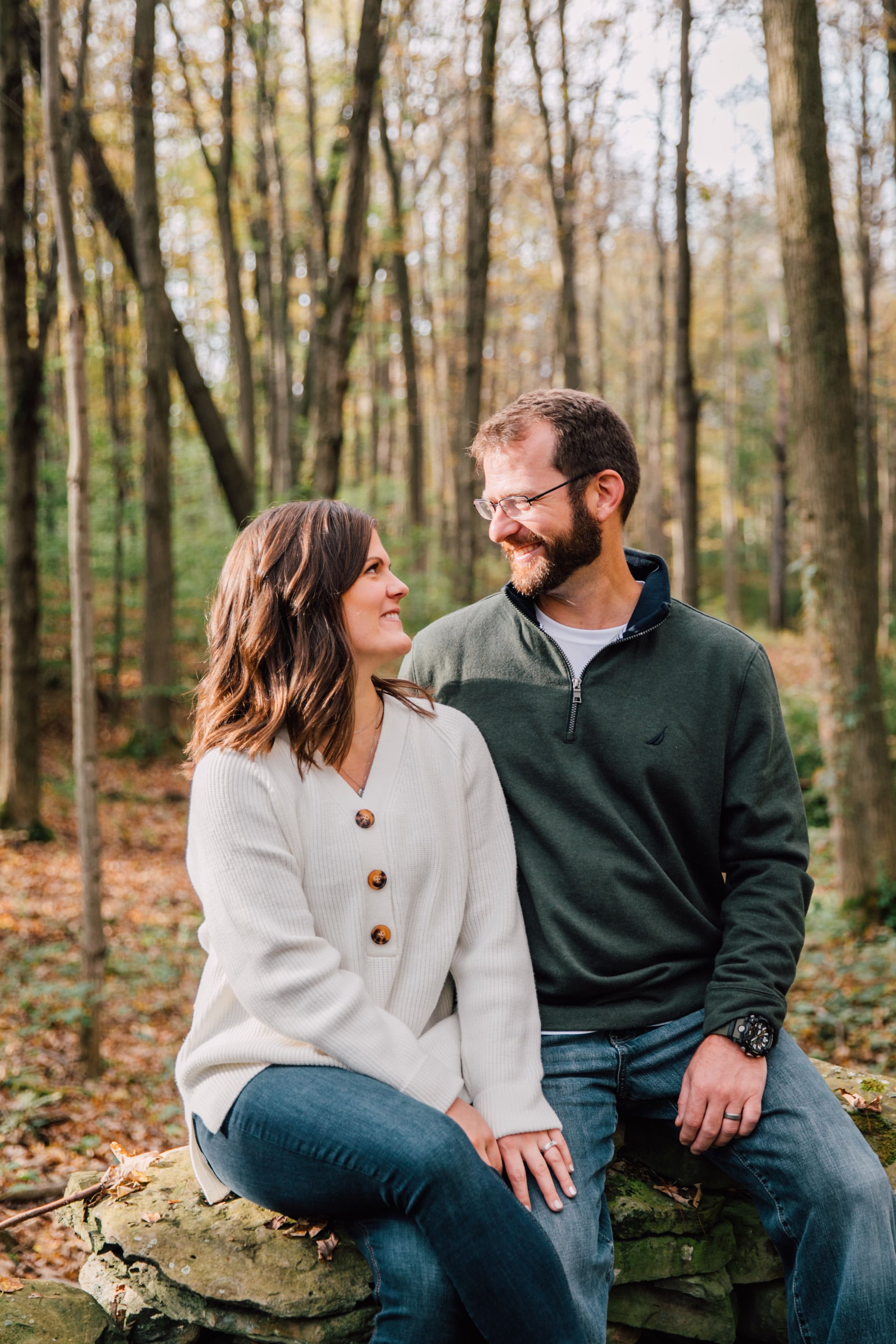  during forest engagement photos, a couple smiles at each other while they sit on a rock wearing fall outfits in a wooded area 