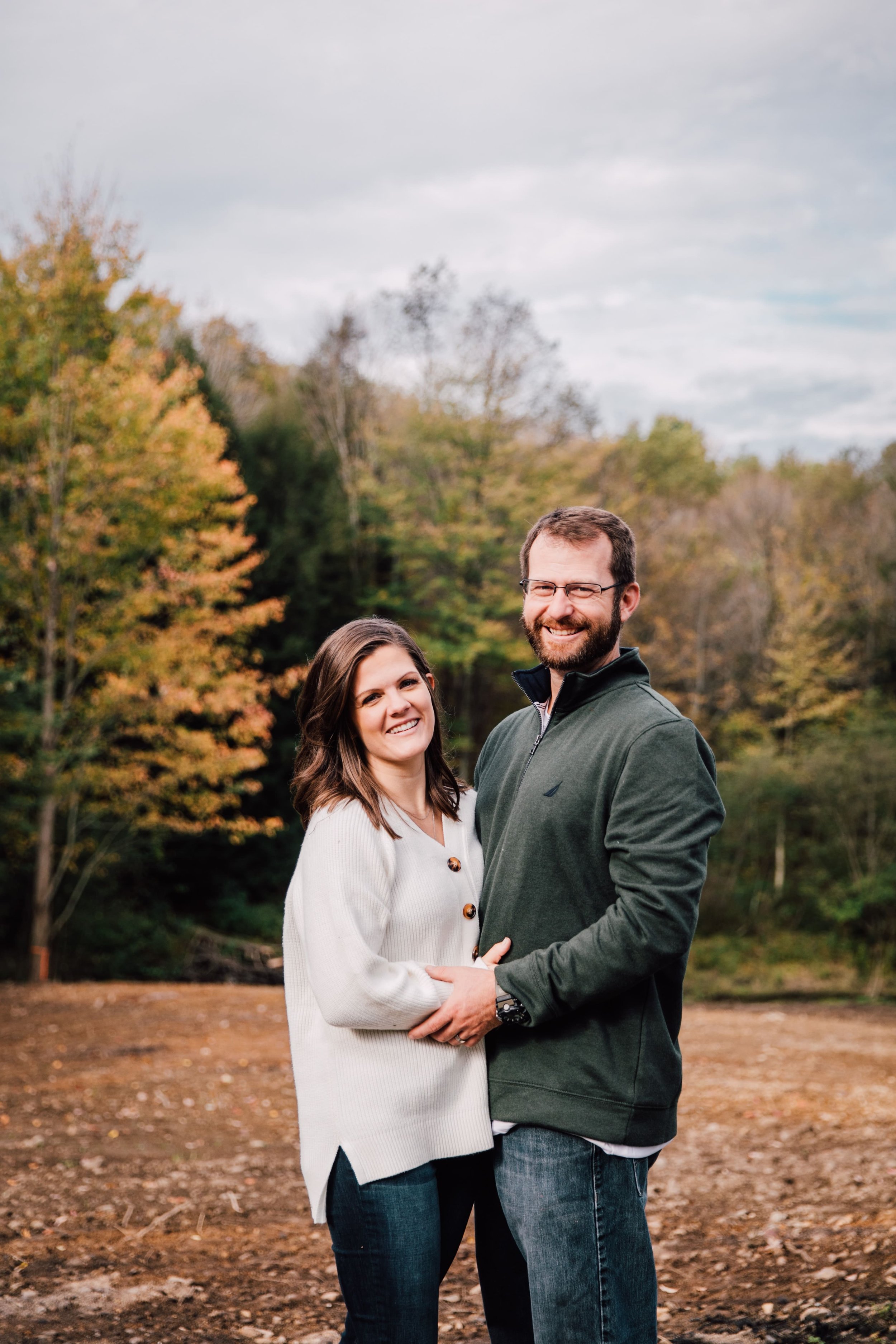  during fall engagement photos a couple stands close together as they smile in front of the wooded area on their property 