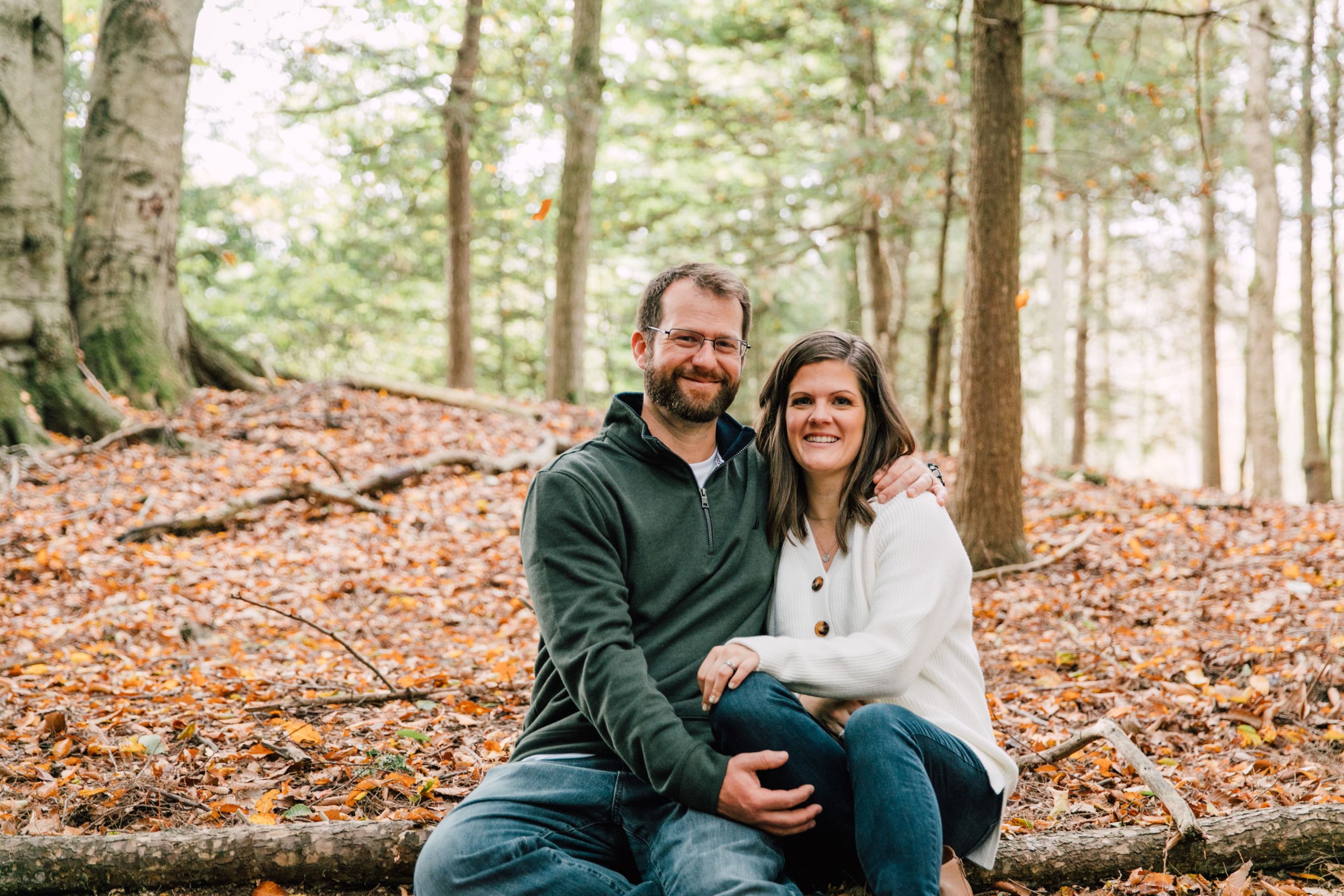  a couple sits together during a couples photoshoot in the fall pictured here they sit on the leaf lined forest floor and smile as they are cuddled up 