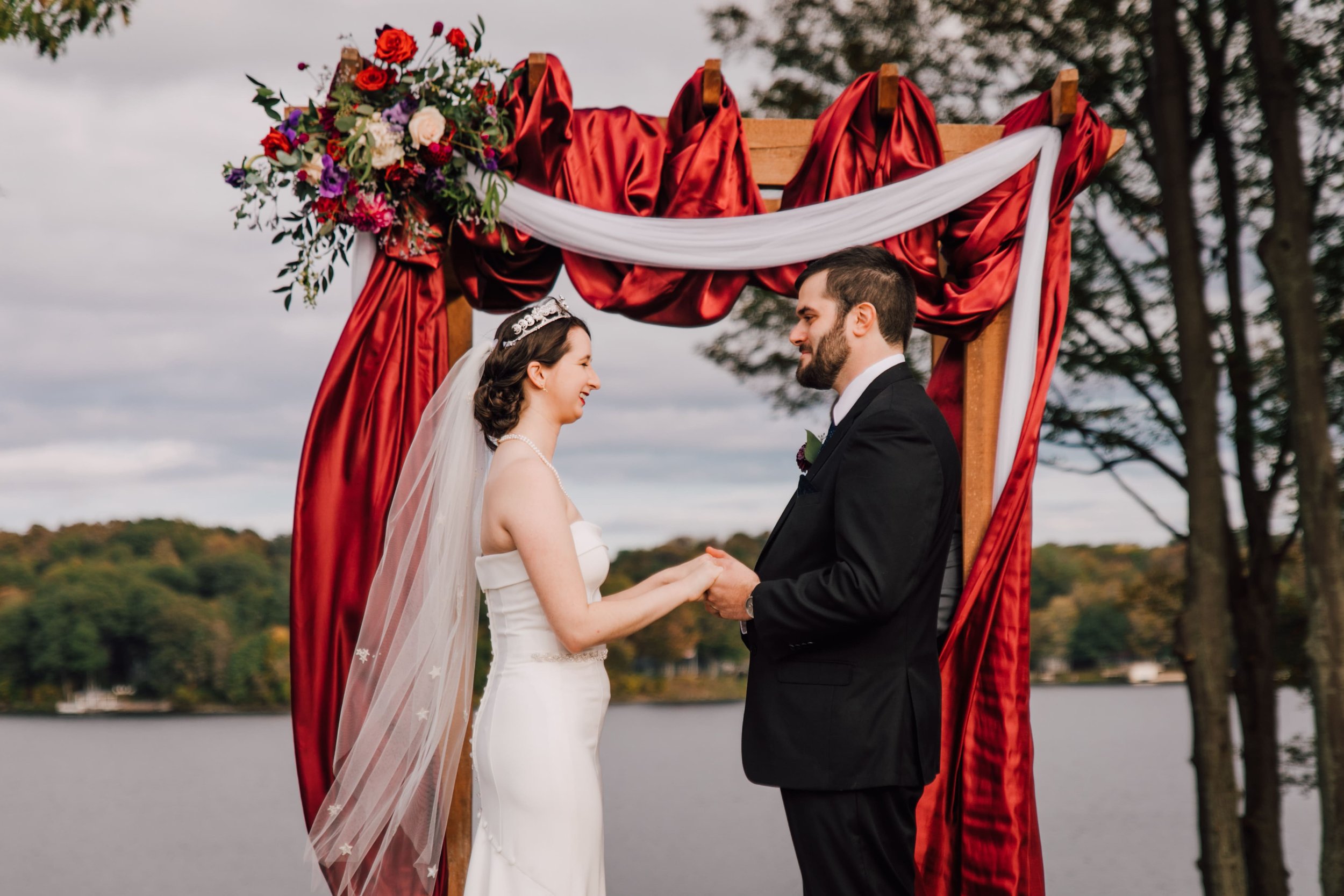  the bride and groom hold hands and smile at each other during their outdoor lake ontario wedding in front of a wedding arbor with red silks and florals 
