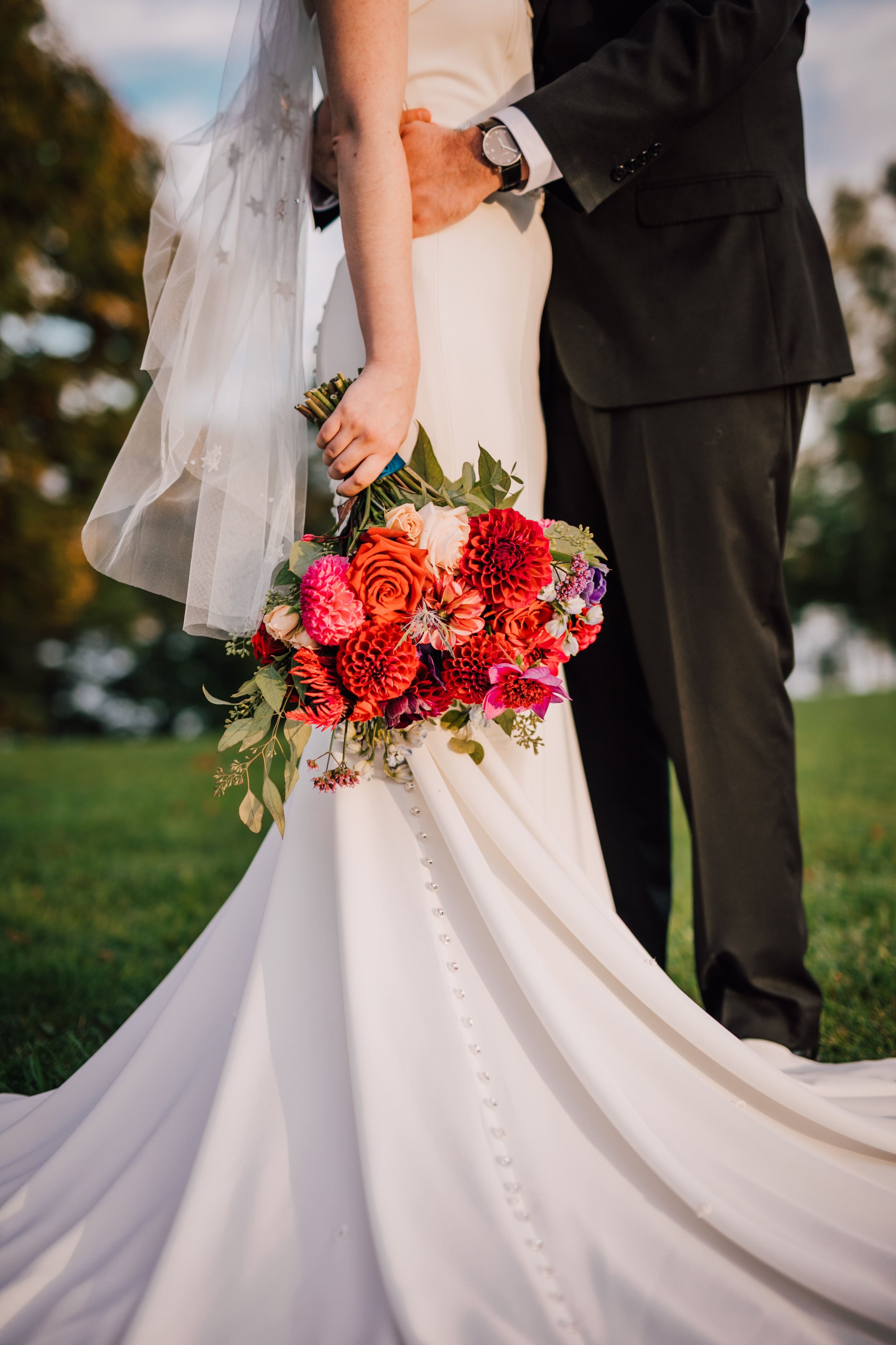  the bride holds her red, pink, and purple bouquet as she stands next to her husband who has his arms wrapped around her waist as she wears a star embroidered veil at their starry night wedding 