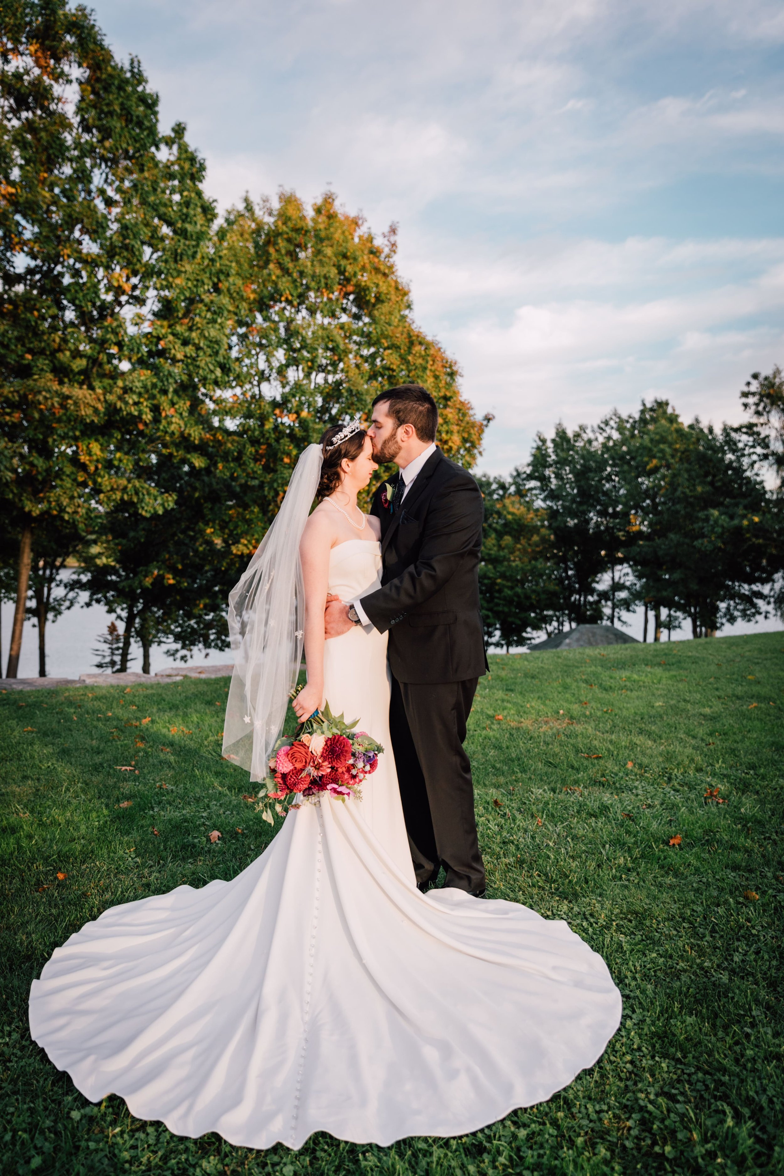  groom stands close to the bride and kisses her forehead as they stand on a lawn with her train sprawled out in front of them at their lake ontario wedding 