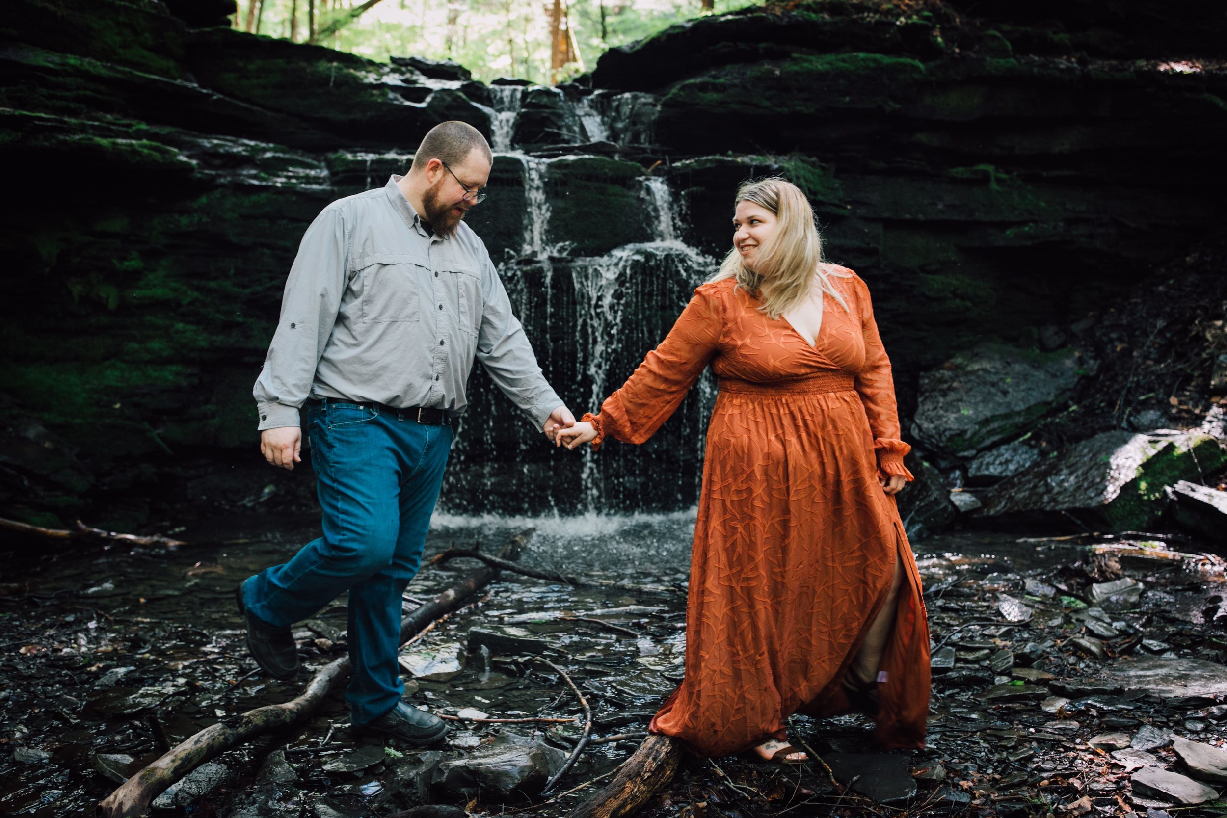  brittany looks back at patrick holding his hand as they walk across a stream while creating their waterfall engagement photos 