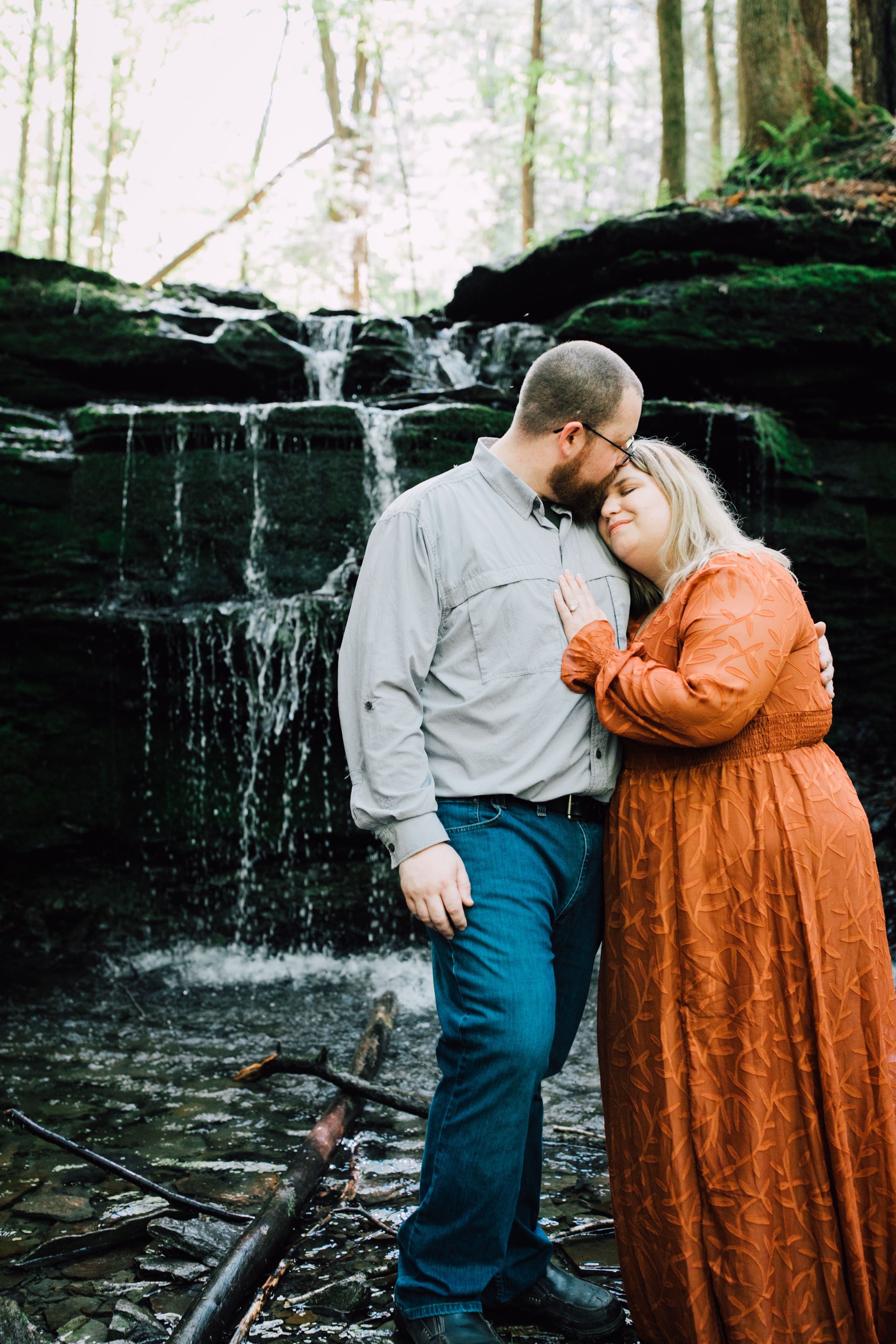  engaged couple stand in front of a waterfall patrick kisses brittany’s forehead during their engagement photos in central new york 