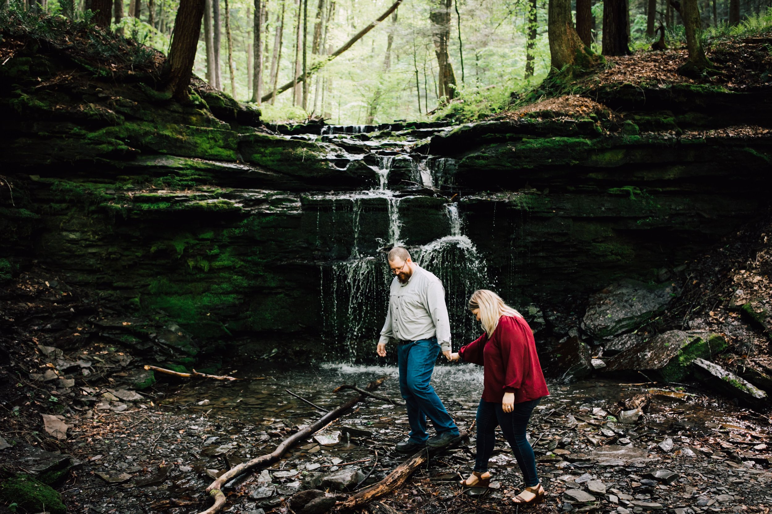  engaged couple hold hands as they walk across a small stream in front of a waterfall at labrador hollow 
