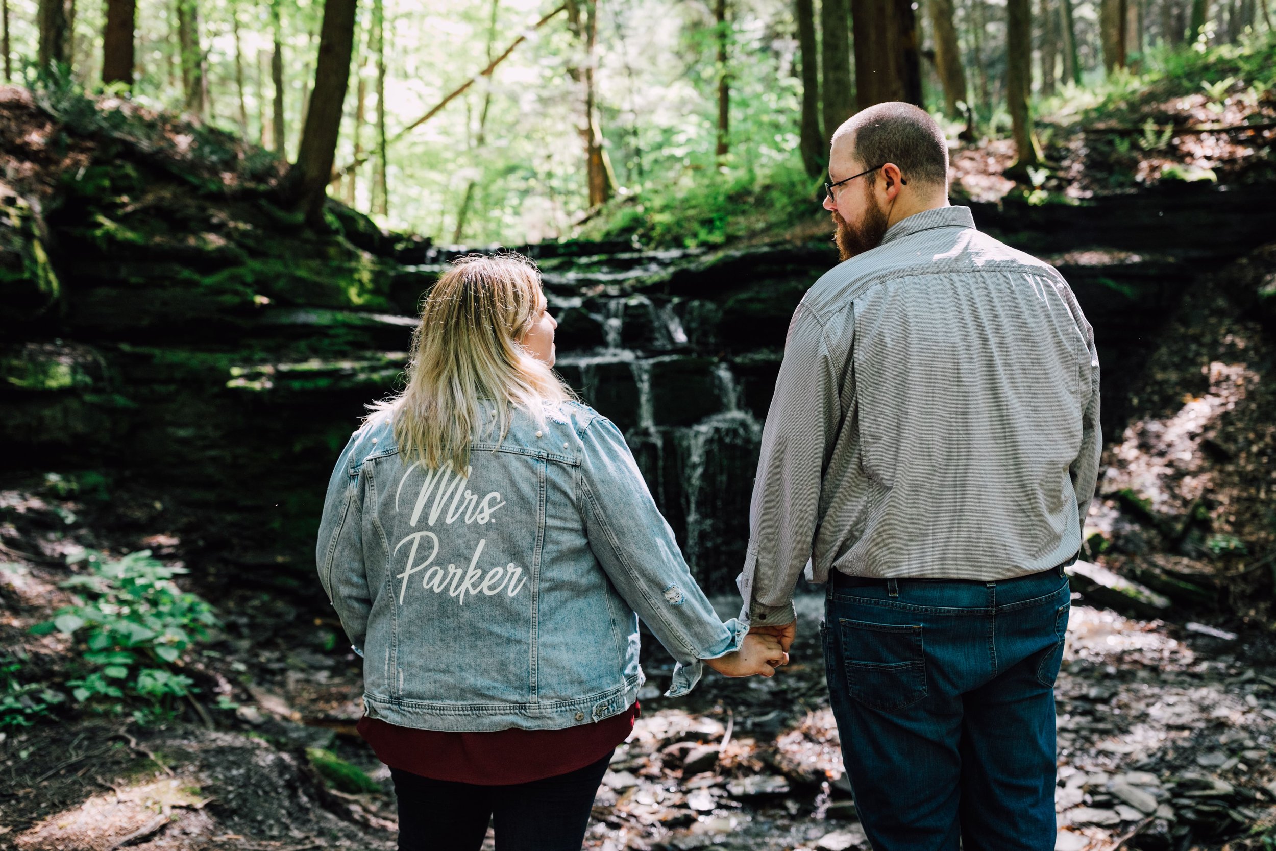  brittany wears her custom wedding jean jacket as she holds her fiancee’s hand as they pose for their forest engagement photos 