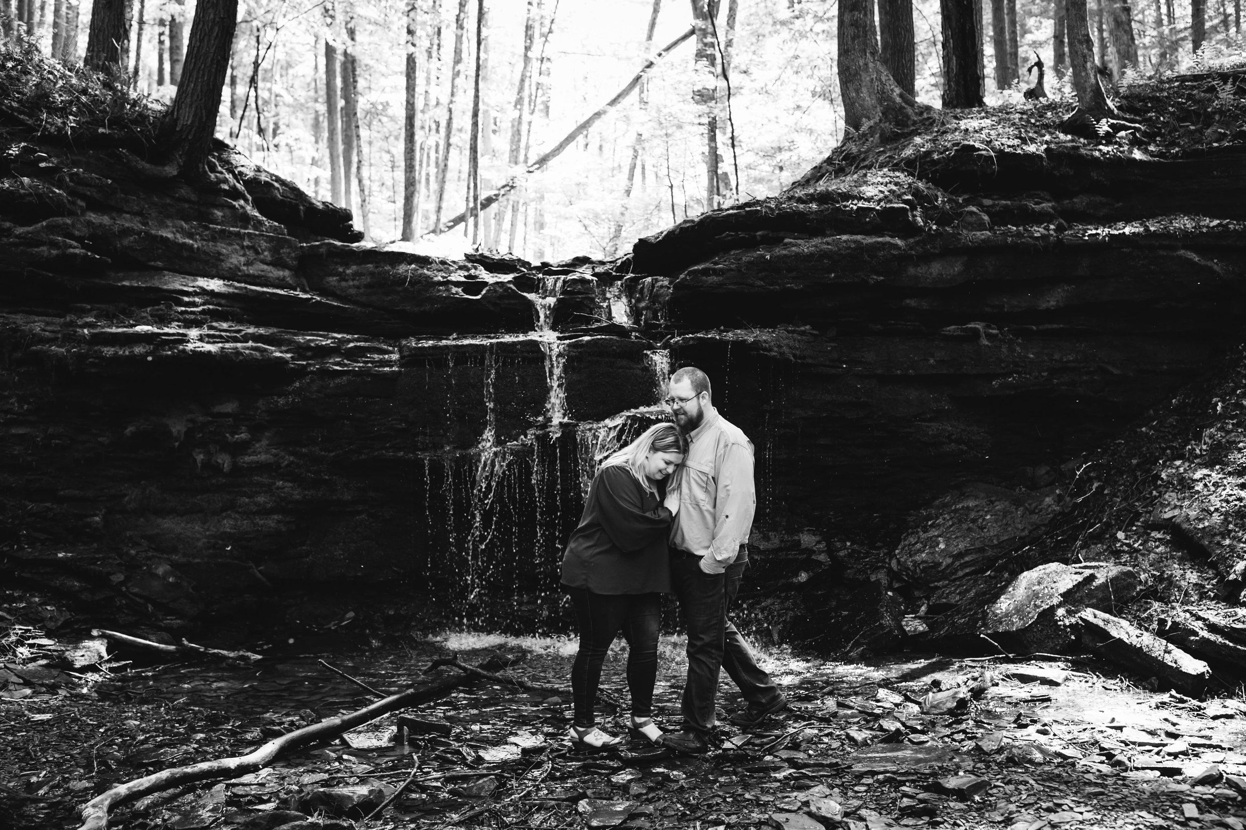  a black and white photo of an engaged couple laughing together as they post for engagement photos in front of a waterfall 