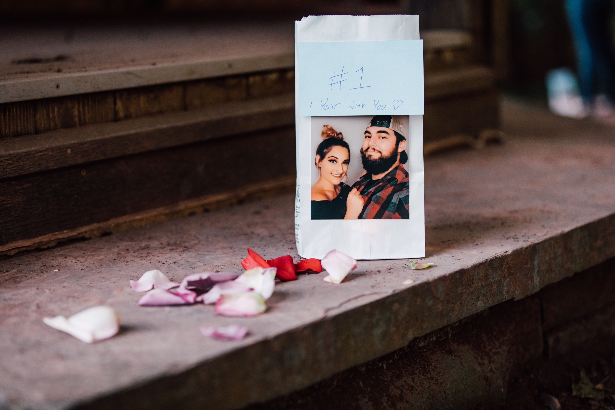  a photo of the engaged couple from their first date is taped to a white paper sack that sits next to flower petals during a proposal at root glen 