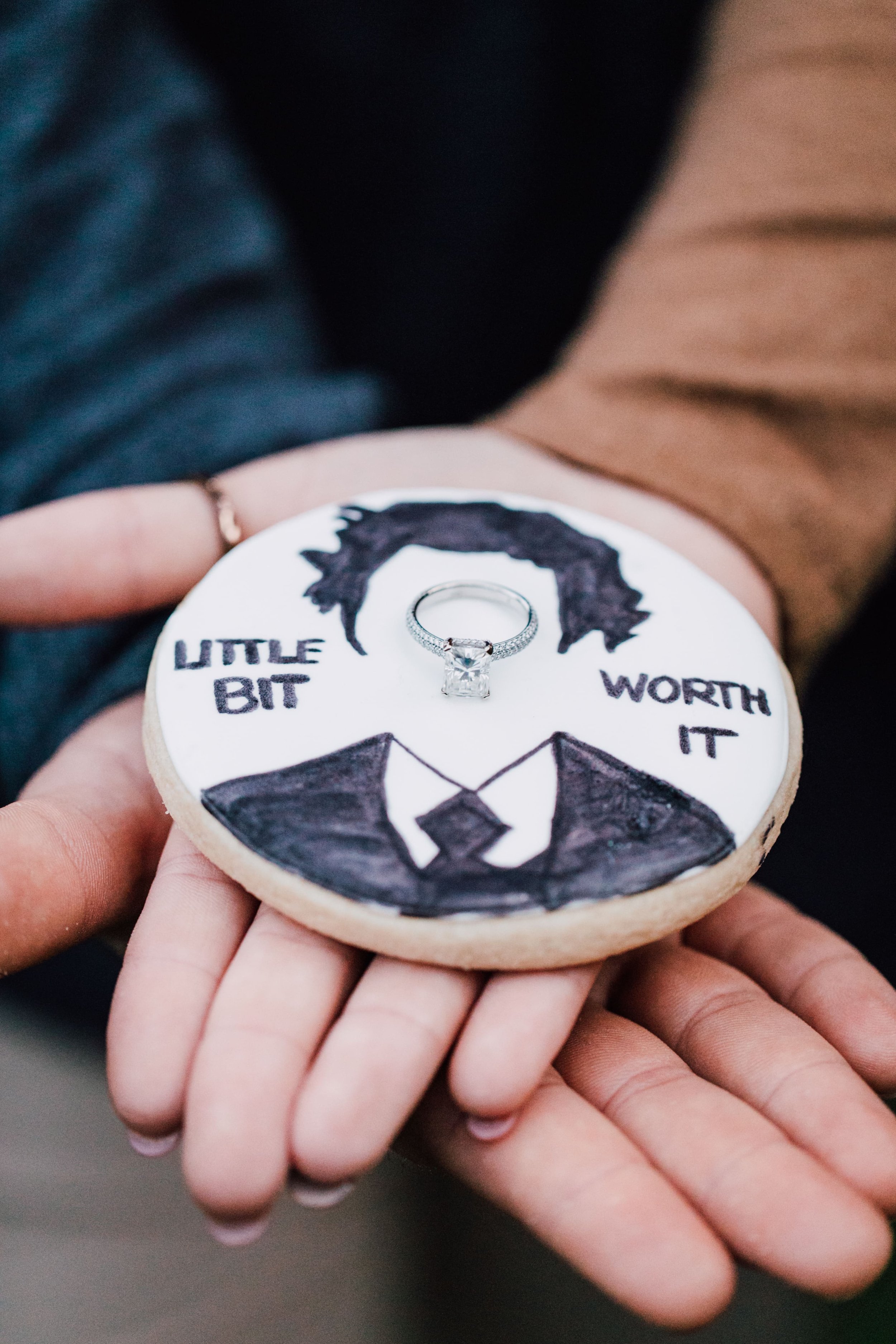  a couple holds their hands out with an engagement ring sitting in the middle of a “the office” themed cookie as proposal photographer captures it all 