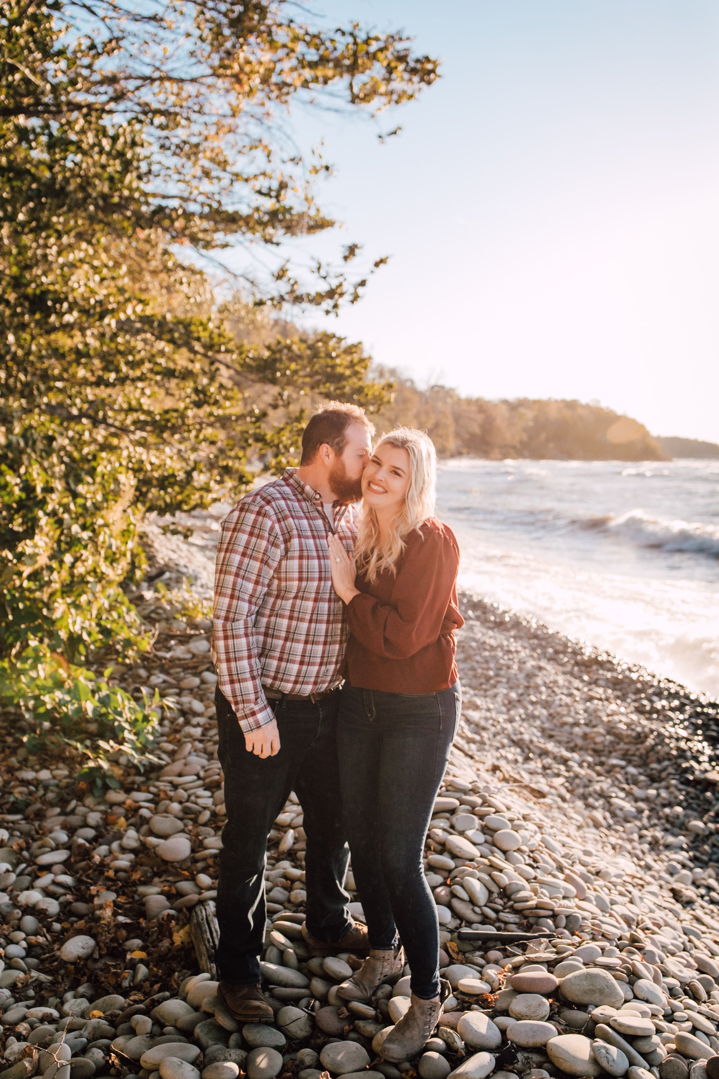  anthony kisses danielle’s cheek as they stand on a rocky beach in front of a great lake for their sunset engagement photos 