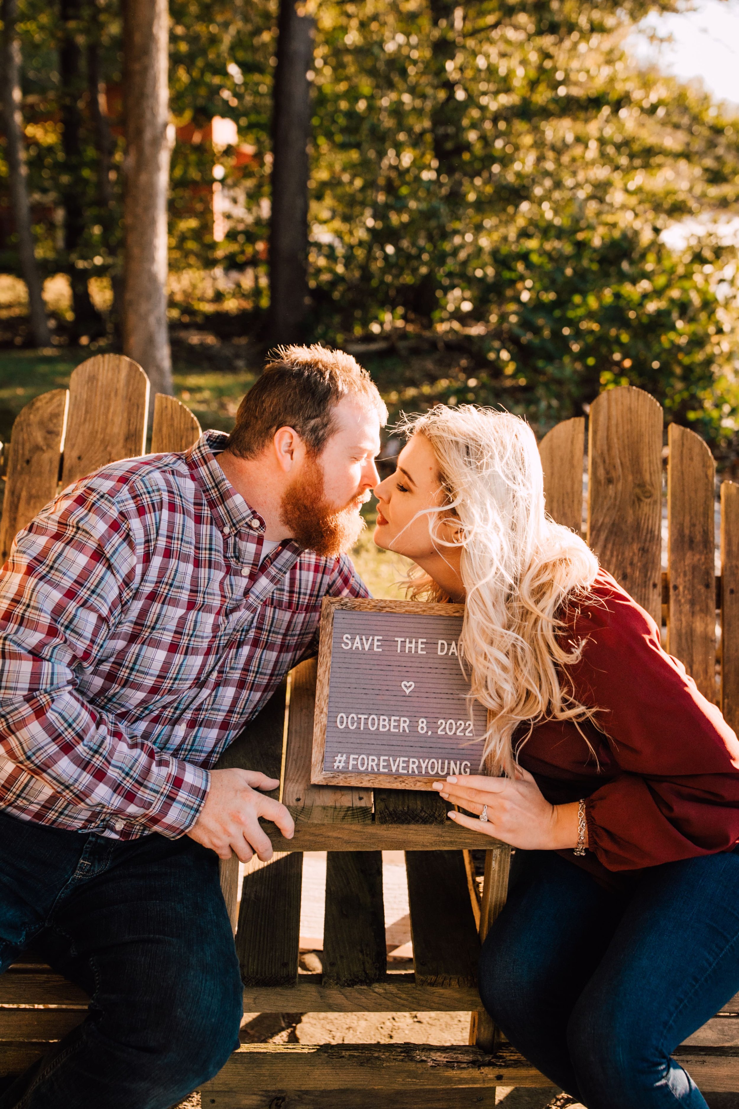  engaged couple lean in for a kiss while they hold a letterboard during their sunset engagement photos that spells out “save the date &lt;3 october 8, 2022 #foreveryoung”  