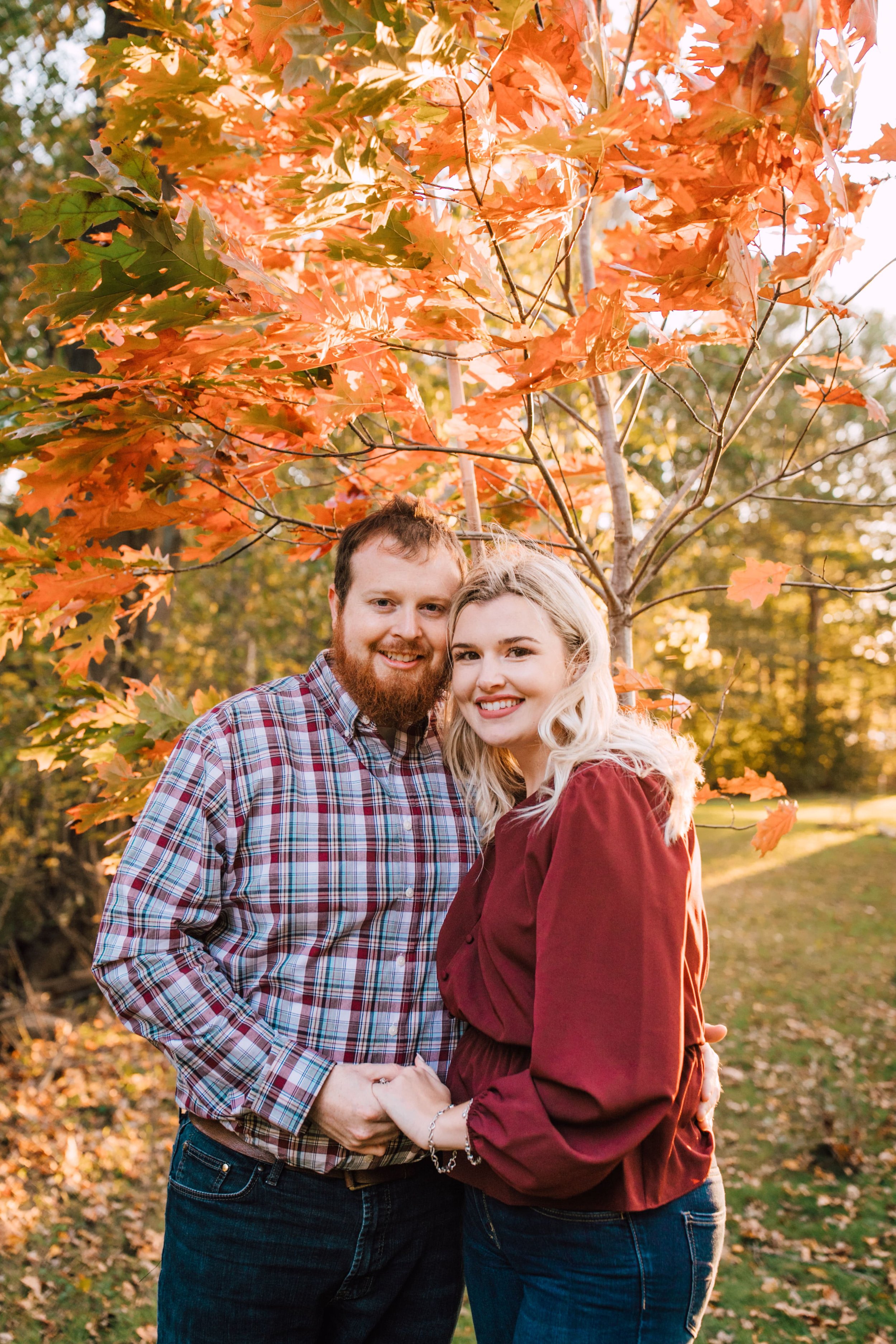  engaged couple stand in front of a tree with changing leaves and smile as the lean against each other and hold hands during the fall engagement photos 