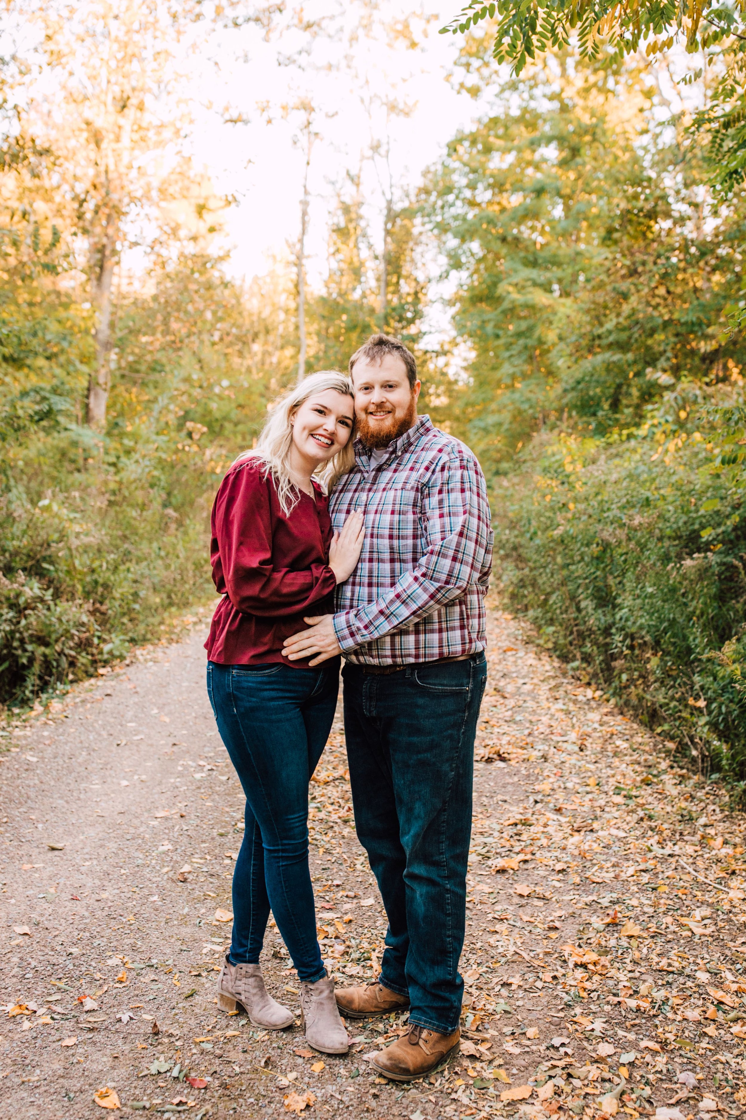  engaged couple smiles as they lean against each other in the middle of a path in a wooded area during their fall engagement photos 