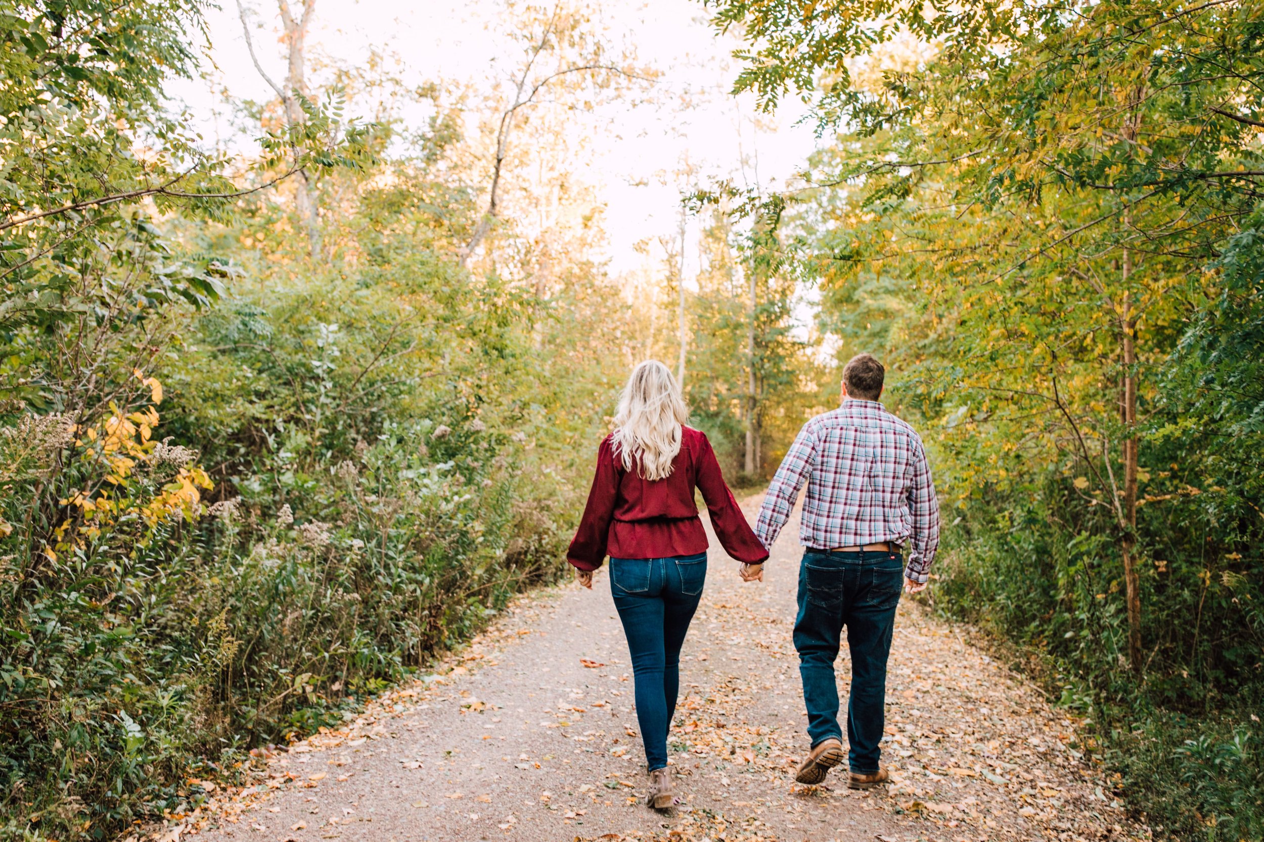  engaged couple holding hands as they walk away from their engagement photographer down a path in a wooded area for their fall engagement photos 