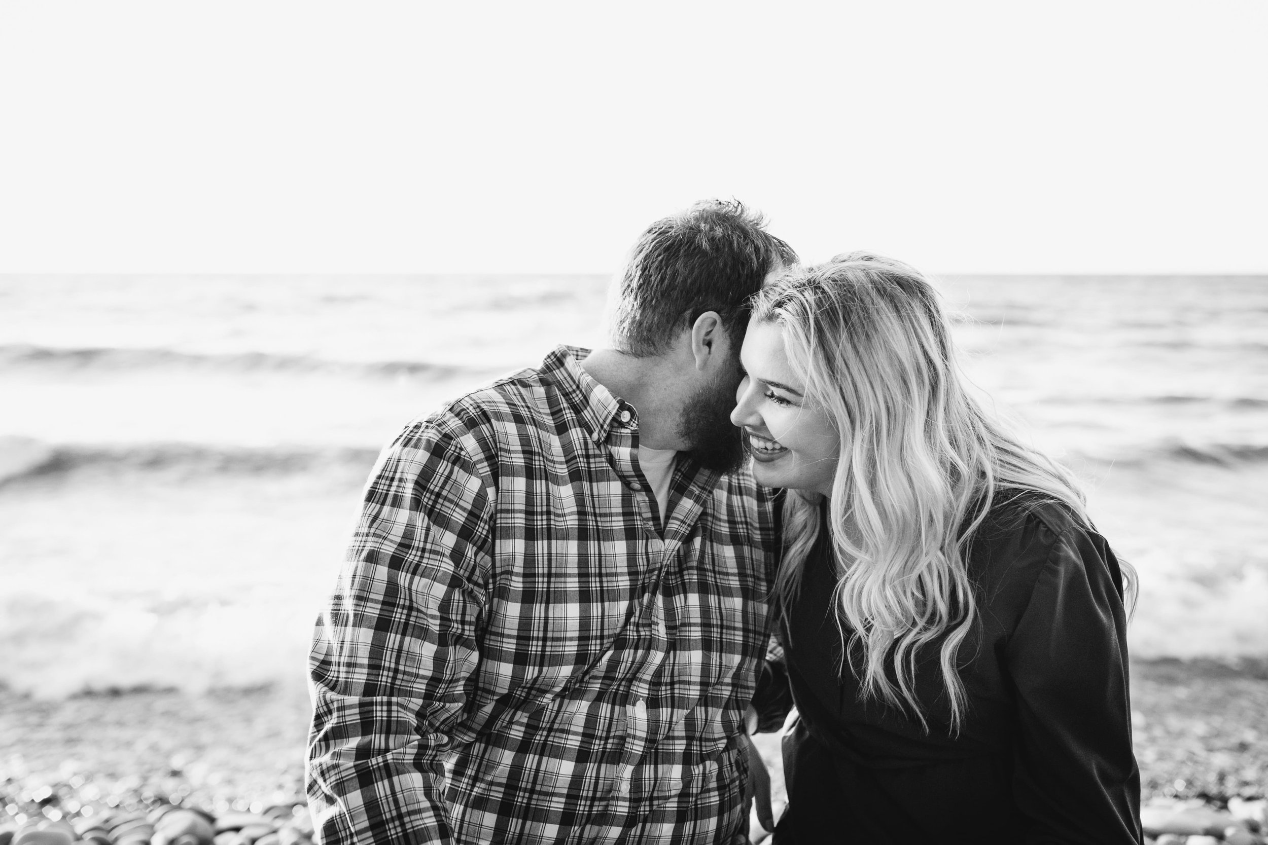  danielle laughs as she presses her cheek to anthony’s face who is seemingly whispering in her ear as they sit in front of a great lake for their lake engagement photos 