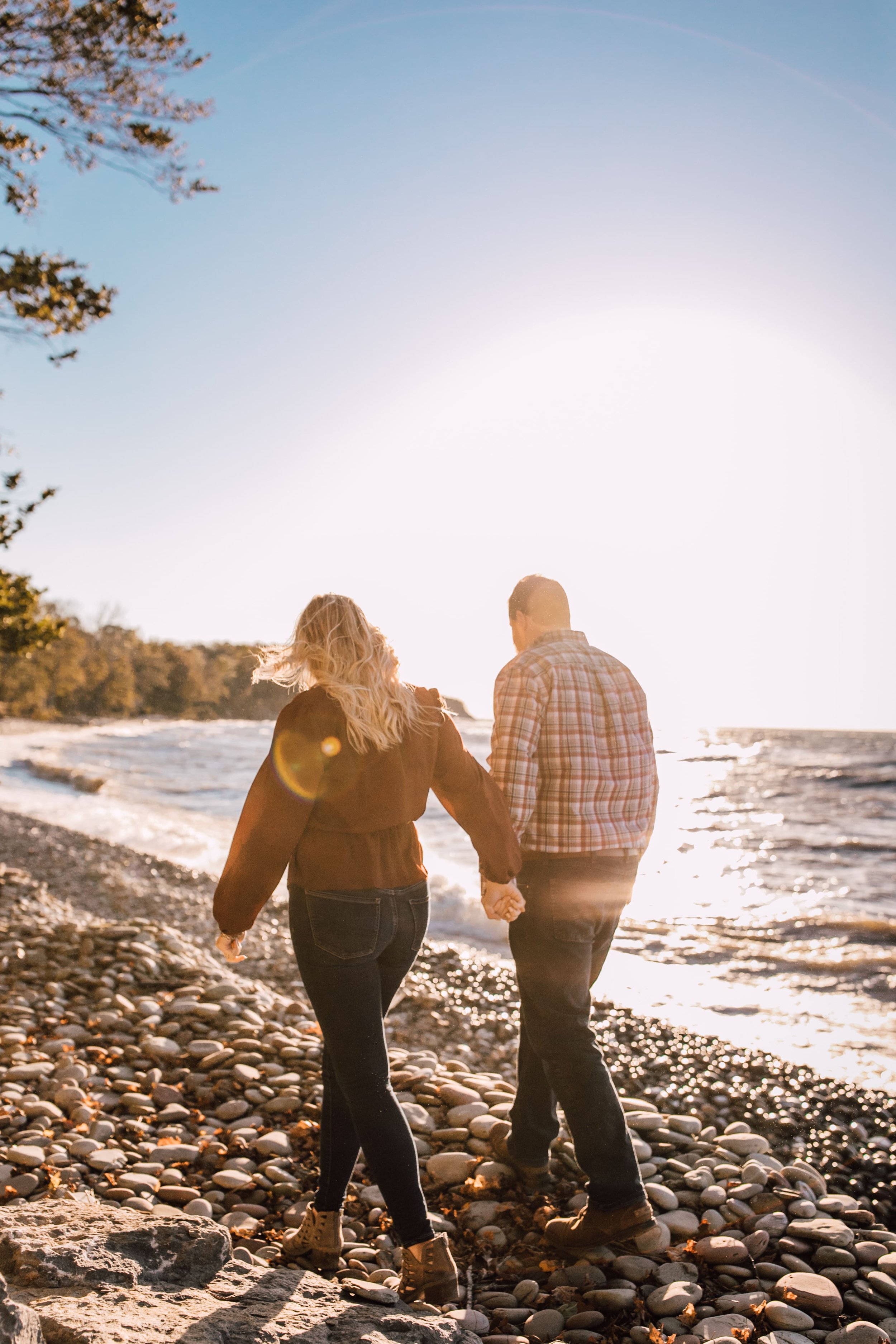  sun flares decorate this photo of an engaged couple walking hand in hand down a rocky beach on the shore of a great lake for their lake engagement photos 