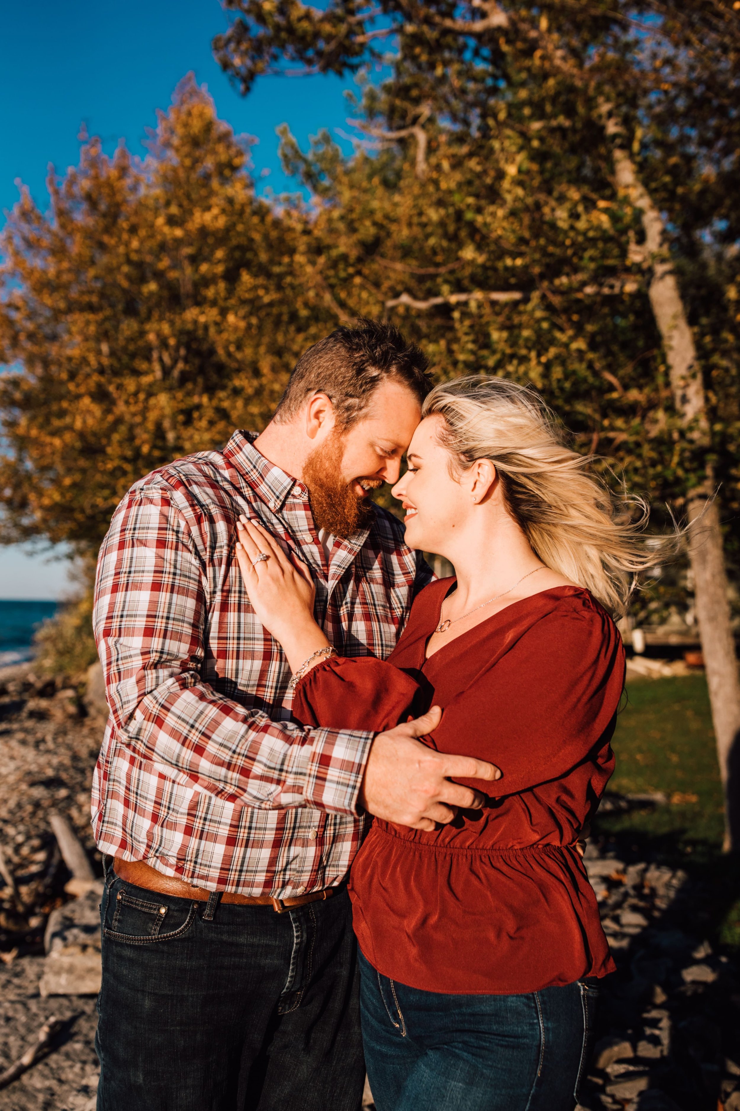  anthony and danielle smile as they stand with their foreheads pressed together for their lake engagement photos in the fall on the rocky shore of a great lake 