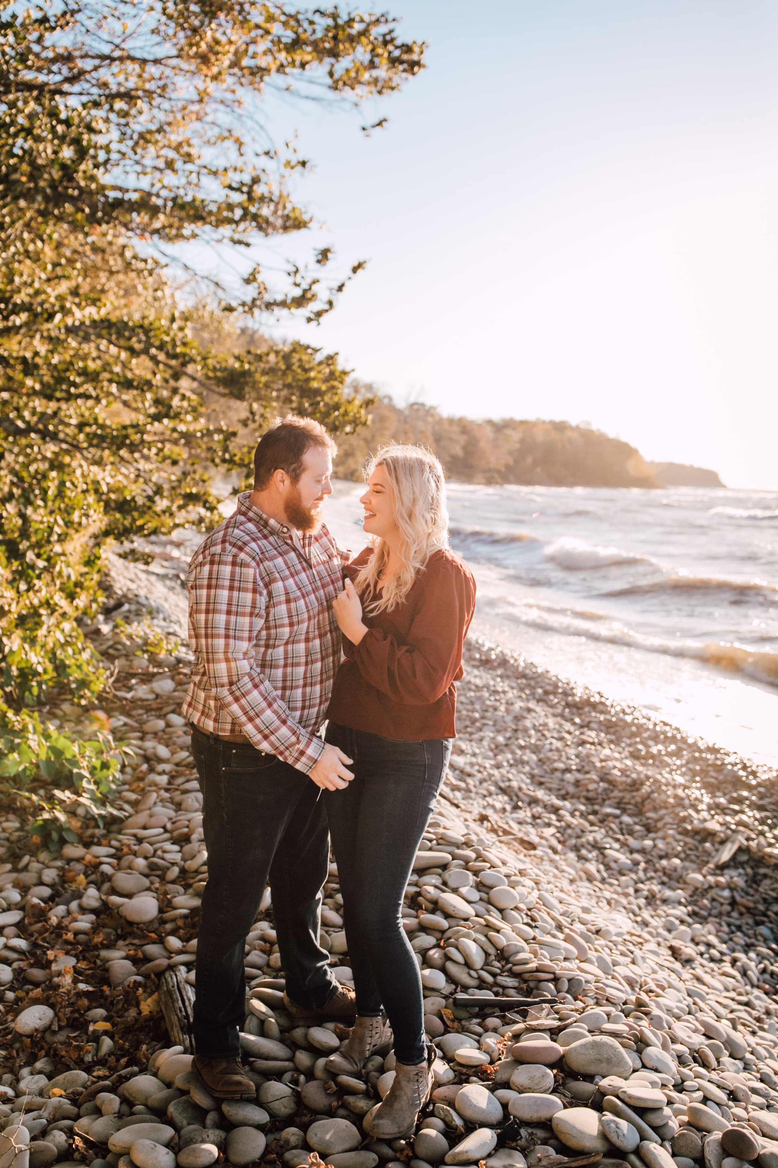  engaged couple smiles at each other as they stand on a rocky lake shoreline for their fall engagement photos 