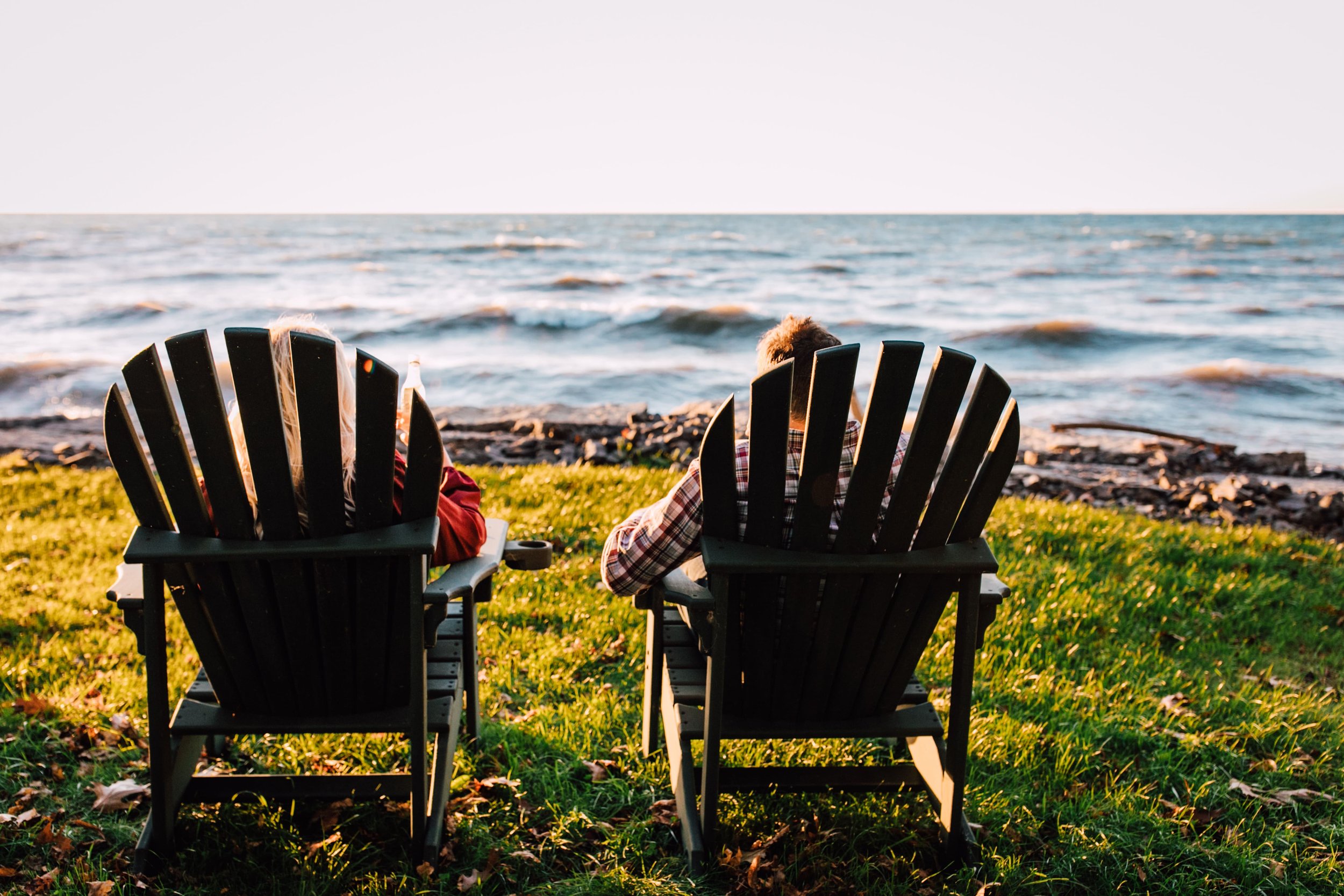  engaged couple enjoy wine coolers while sitting in adirondack chairs facing the rocky shoreline of a great lake as the sun sets on their sunset engagement photos 