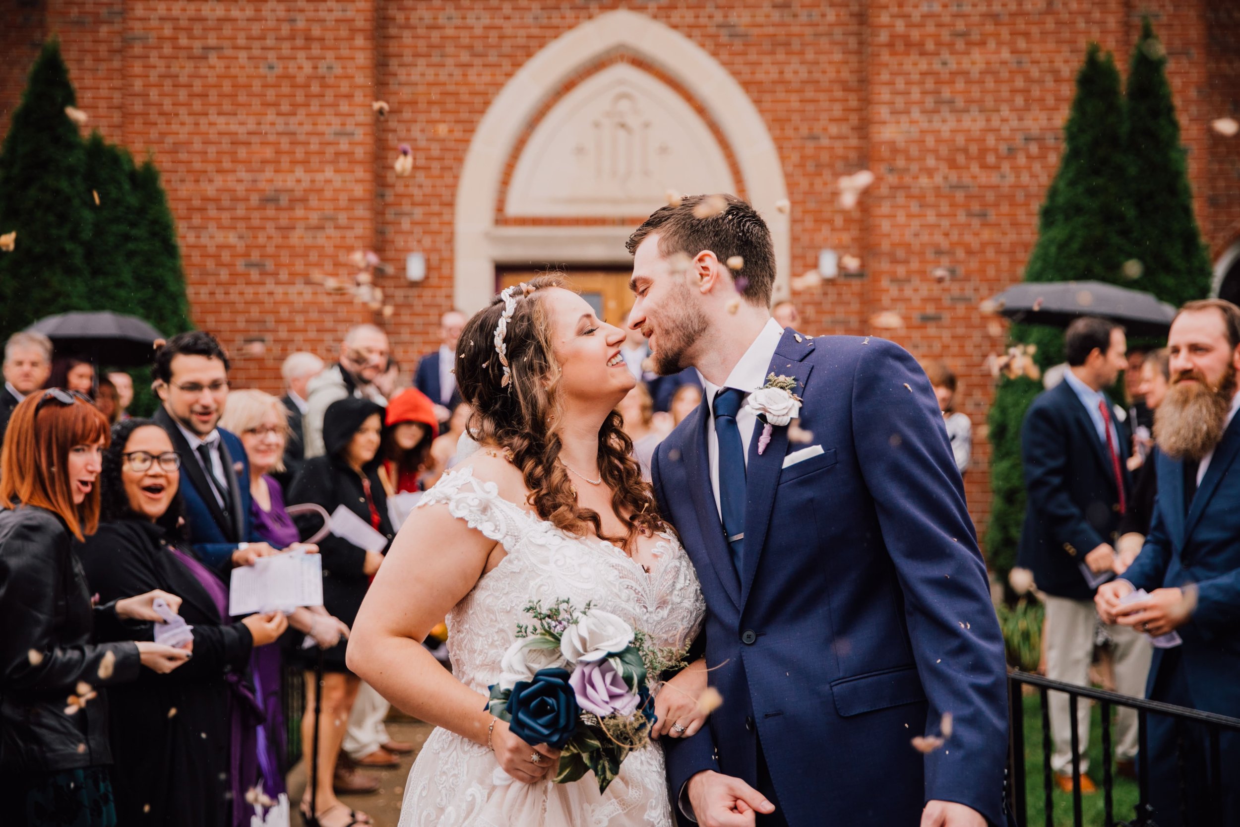  the bride and groom lean in for a kiss outside of the church while their wedding guests throw dried florals and celebrate with them the bride holds a bouquet of paper wedding flowers while the groom wears a paper wedding flower boutonniere  