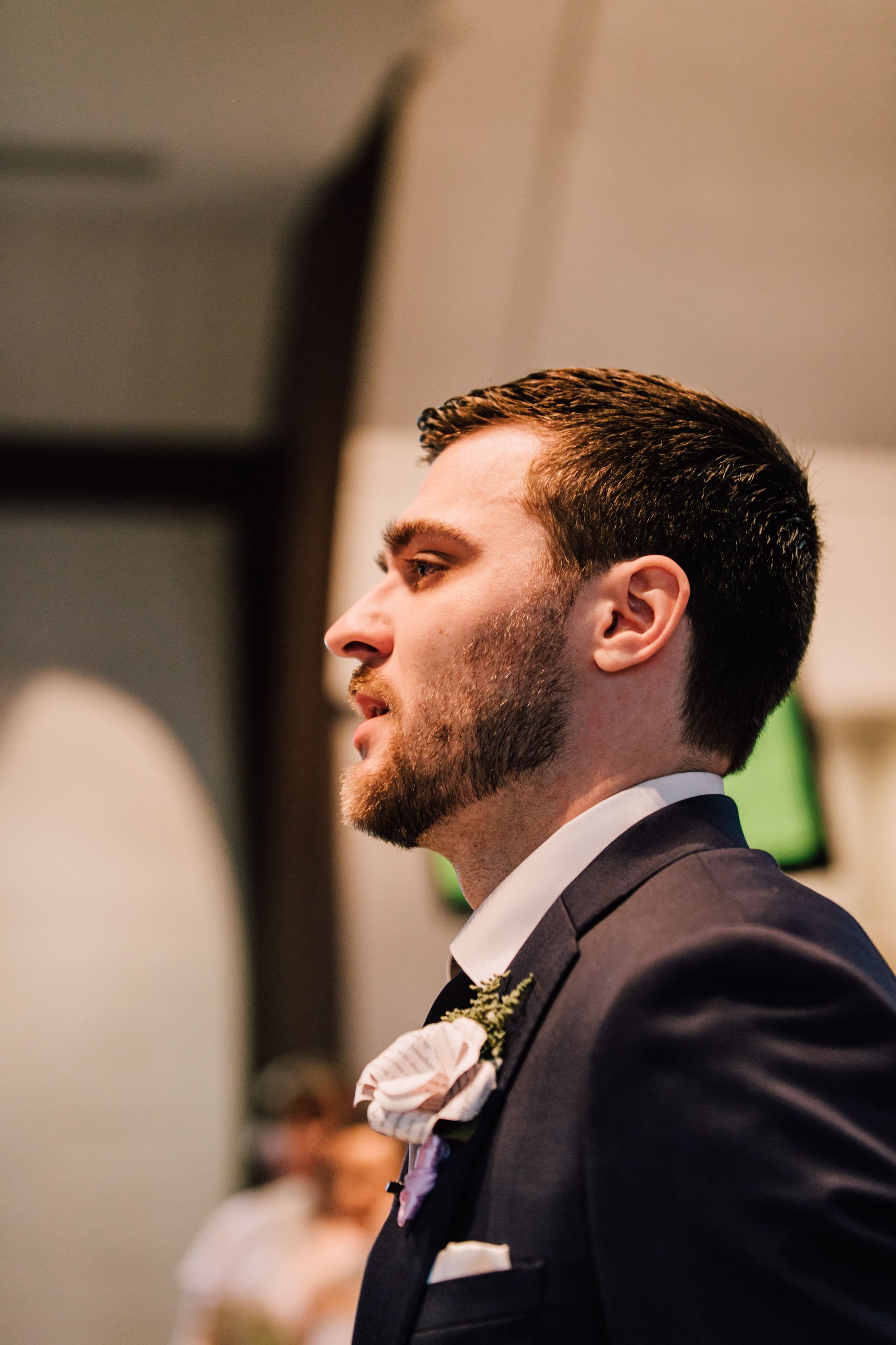  the groom looks adoringly at his fiancee walking down the aisle as he stands on the altar wearing his paper wedding flowers boutonnière   