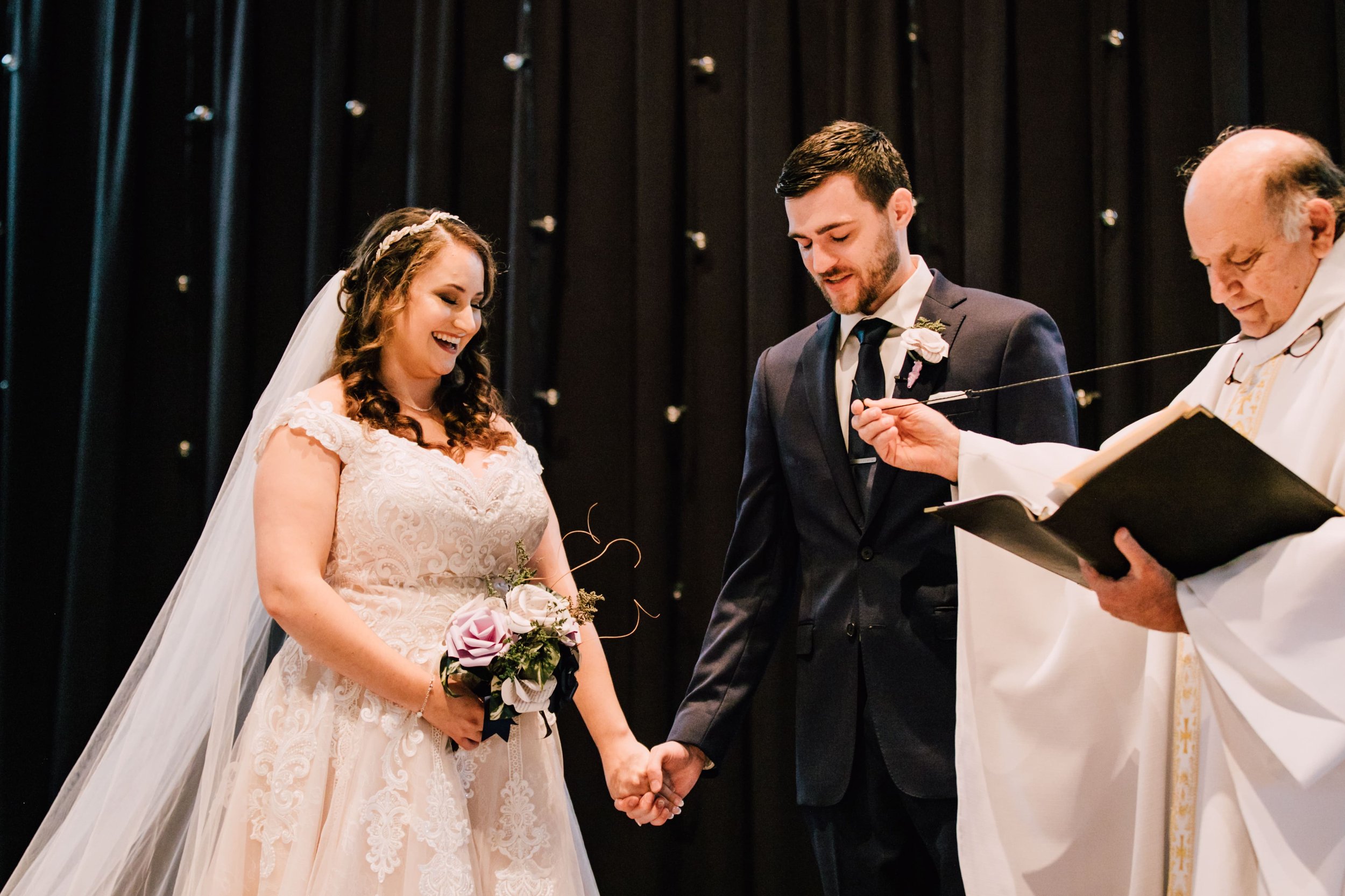  the bride and groom stand hand in hand as their officiant holds out his microphone for them to read their vows. the bride is holding her bouquet of paper wedding flowers  